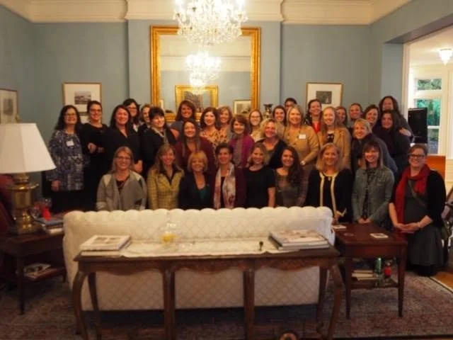 Group of women gathered in a nicely decorated room for a social event or meeting, with a large mirror, chandelier, and framed pictures on the walls.