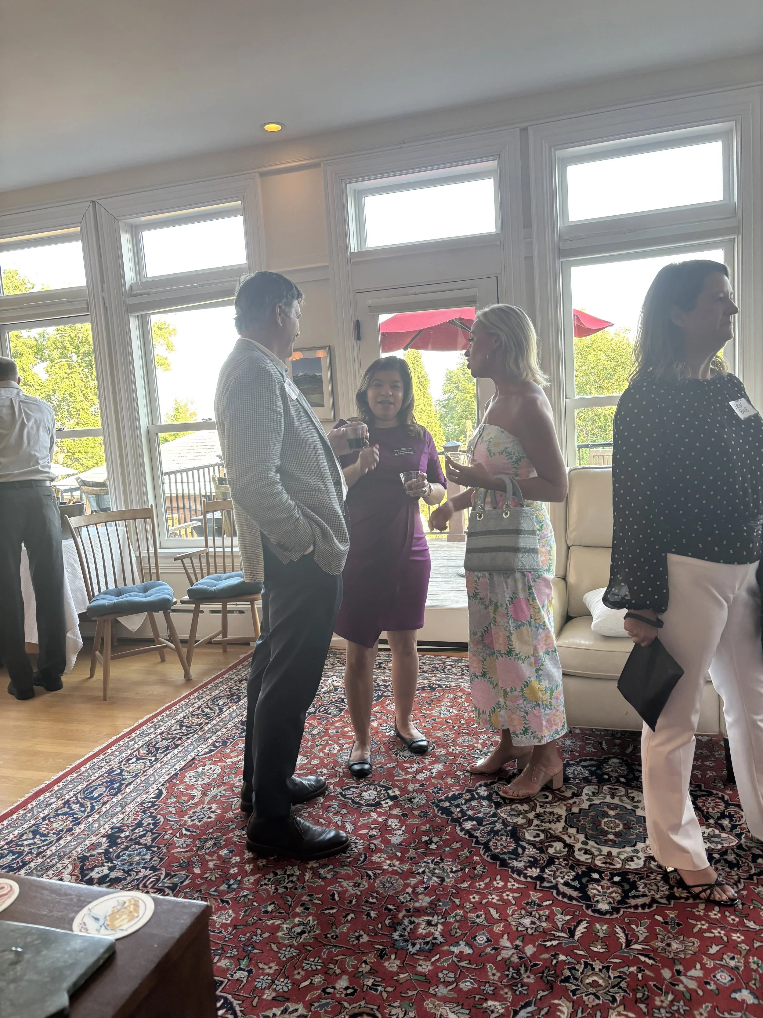 Four women and one man standing and engaging in conversation indoors during a social event, with large windows revealing trees and patio umbrellas outside. The group includes a woman in a floral dress, a woman in a purple dress, a man in a checked bl
