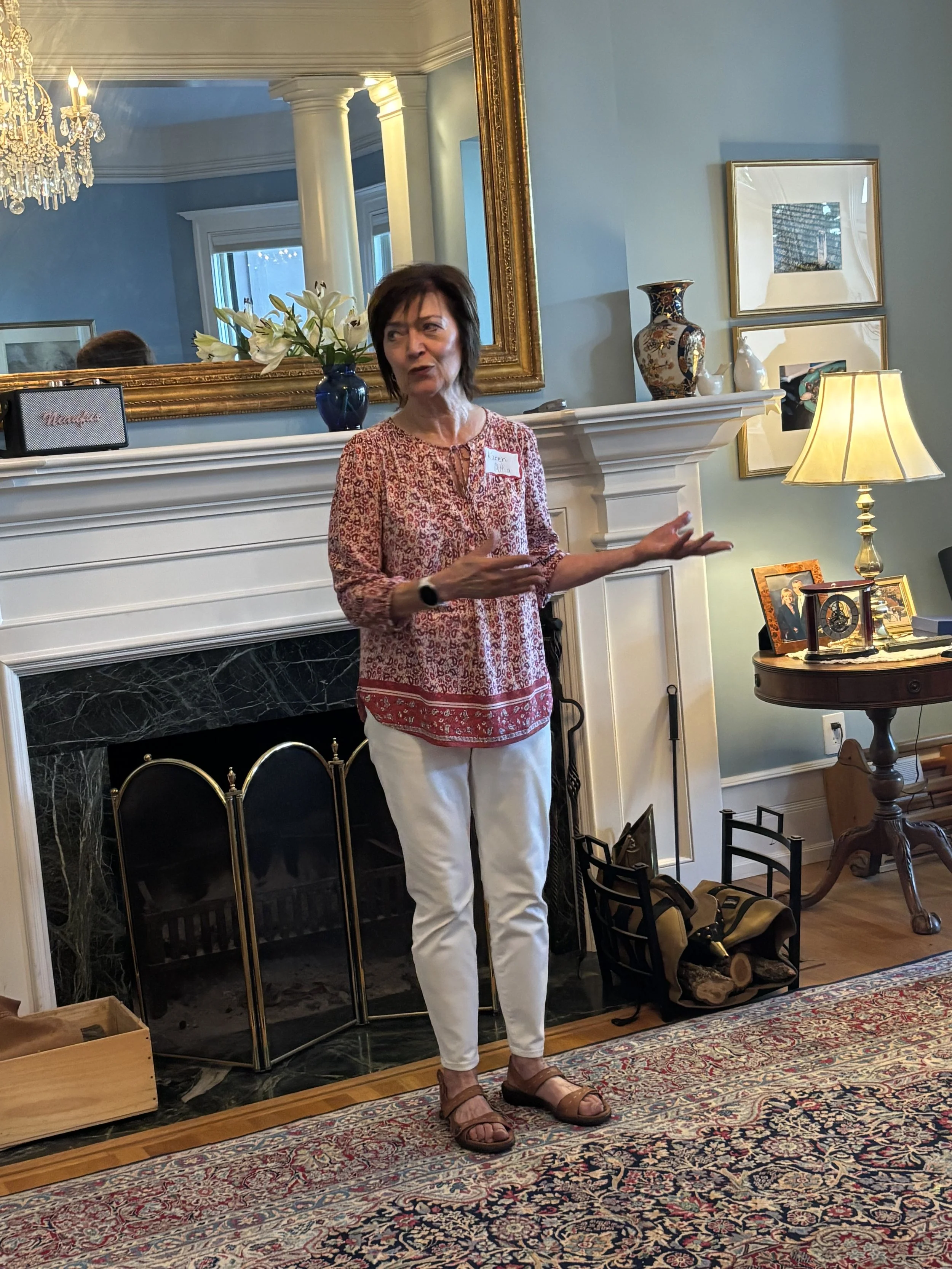 A woman with short brown hair wearing a patterned red and white blouse, white pants, and sandals is standing in a living room near a fireplace, gesturing with her right hand while speaking.