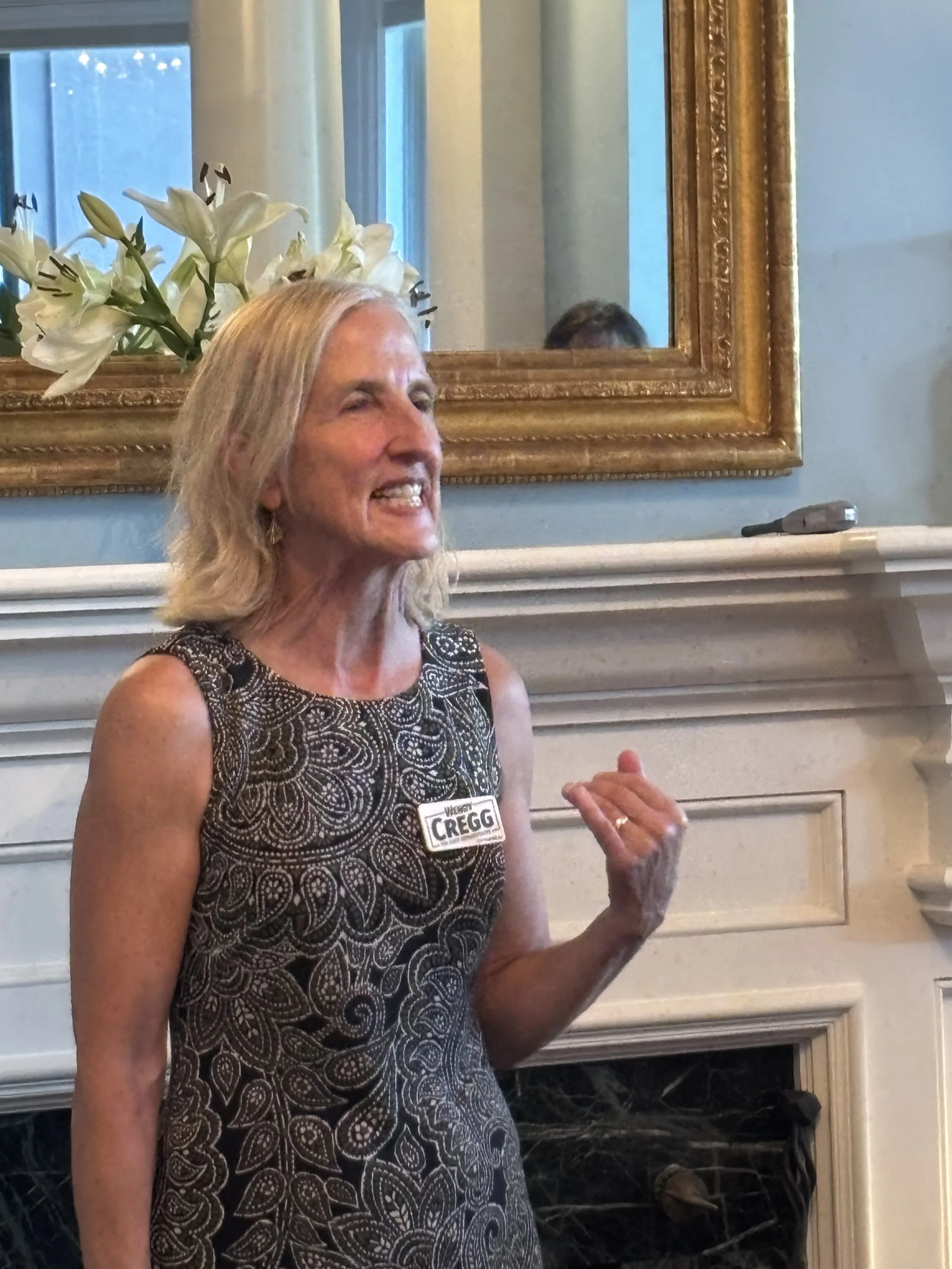 A middle-aged woman with long blonde hair smiling and speaking at an indoor event, wearing a sleeveless black and white patterned dress and a name tag. Behind her is a fireplace with a white mantel, a large mirror with a wooden frame, and a floral ar