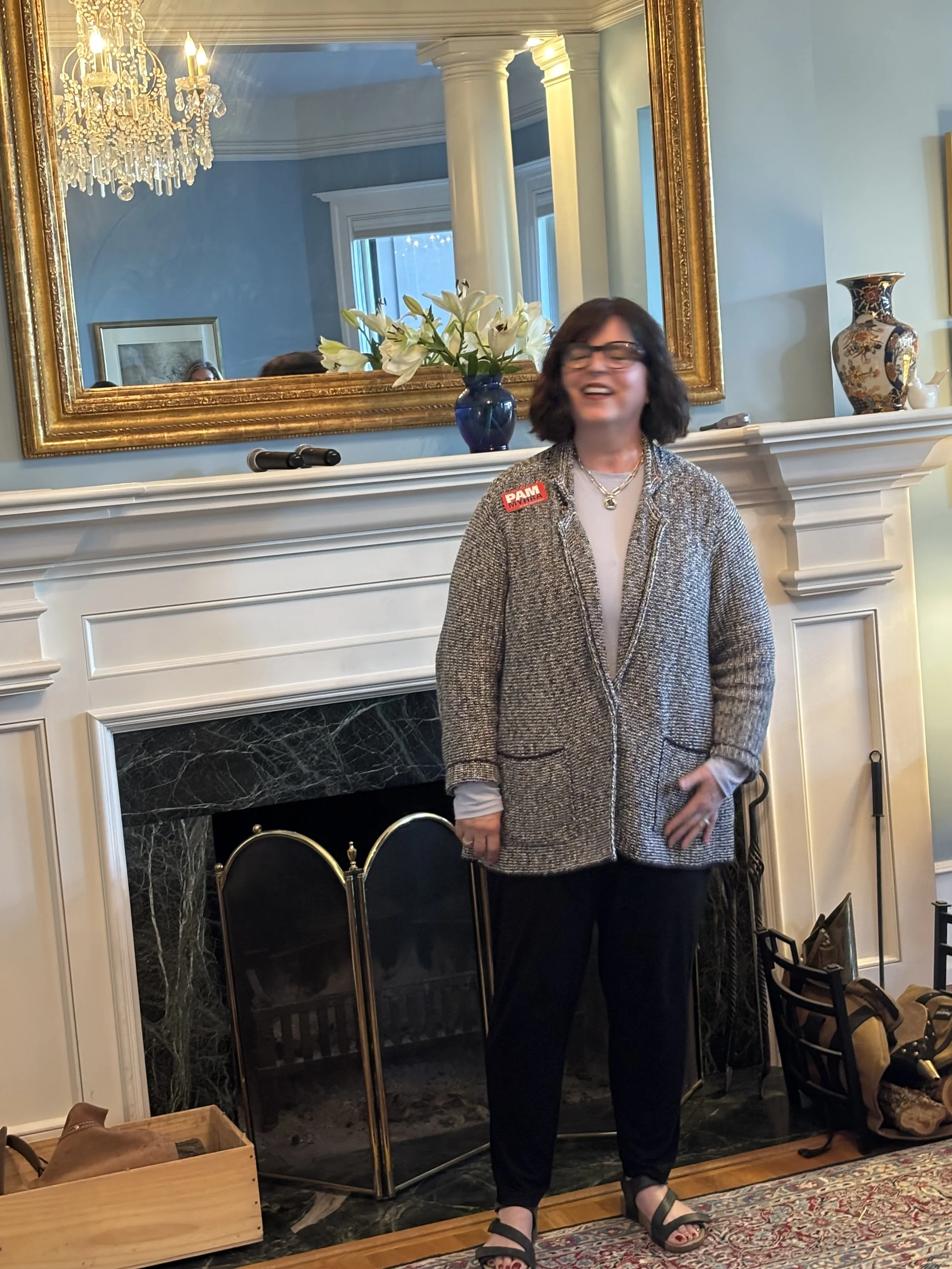 A woman standing in front of a fireplace with a large gold-framed mirror behind her. She is smiling and wearing a gray textured cardigan, black pants, and black sandals. There is a vase of white lilies on the mantel and decorative items nearby.