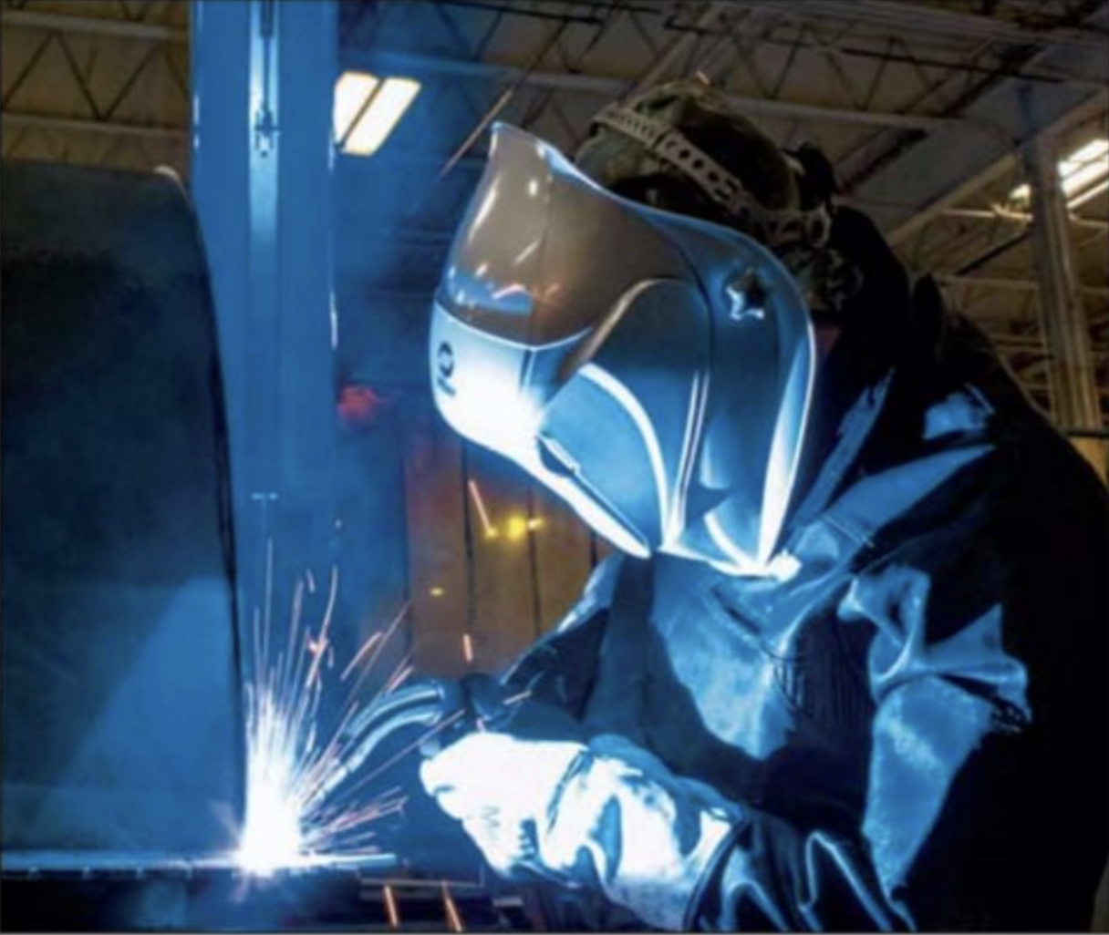 A person welding metal on a manufacturing assembly line, wearing a protective helmet and a dark protective suit, with sparks flying from the welding process.