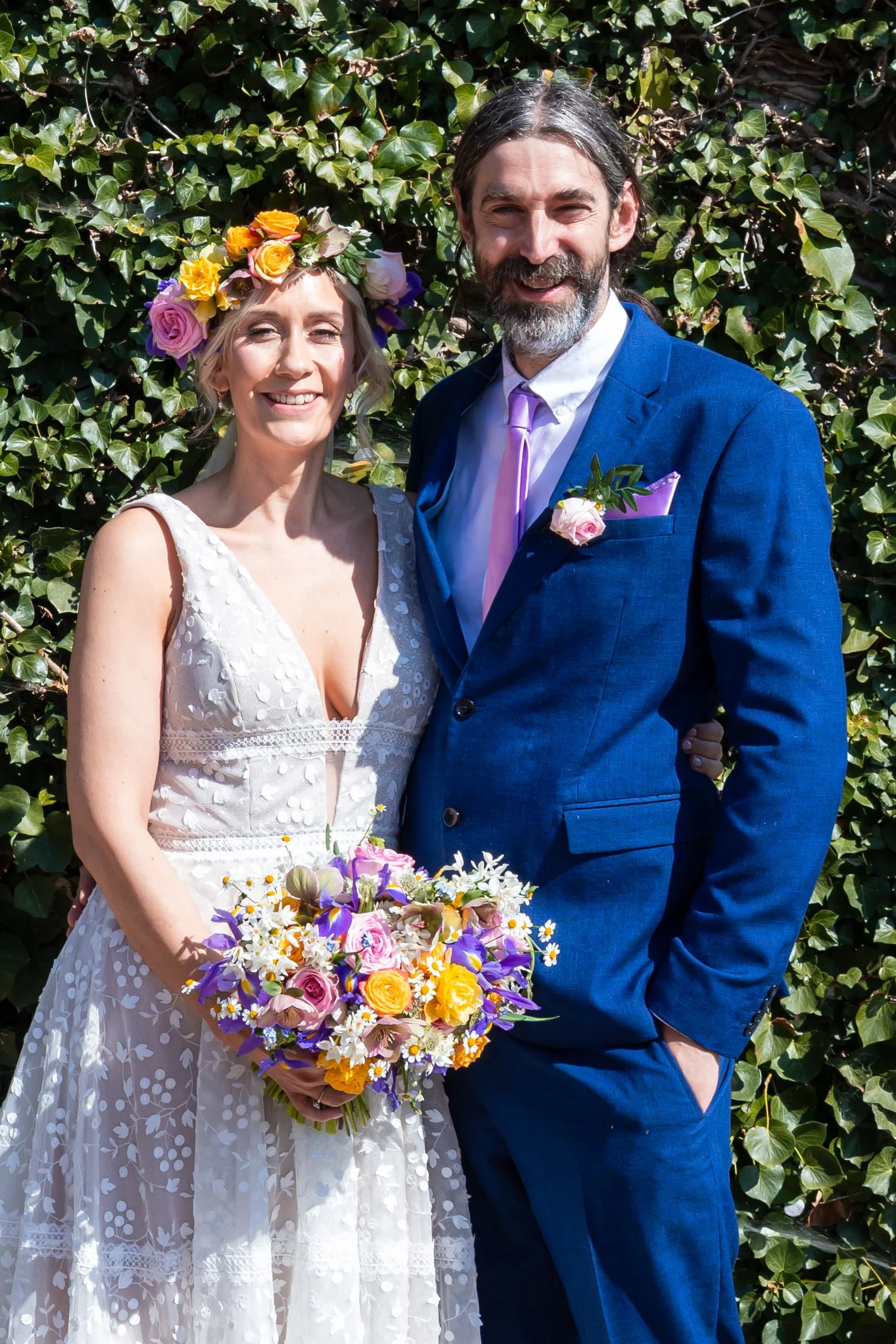 the bride and groom are smiling outside at their Rosedew farm wedding. The bride is wearing a bridal flower crown full of seasonal spring flowers and carrying a gorgeous spring wedding bouquet of colourful spring flowers all made by Petal Power