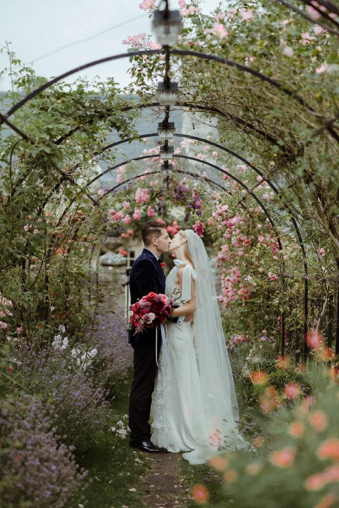 The bride and groom are kissing in a rose garden the bride is holding her bridal flowers in burgundy, red and pink colours