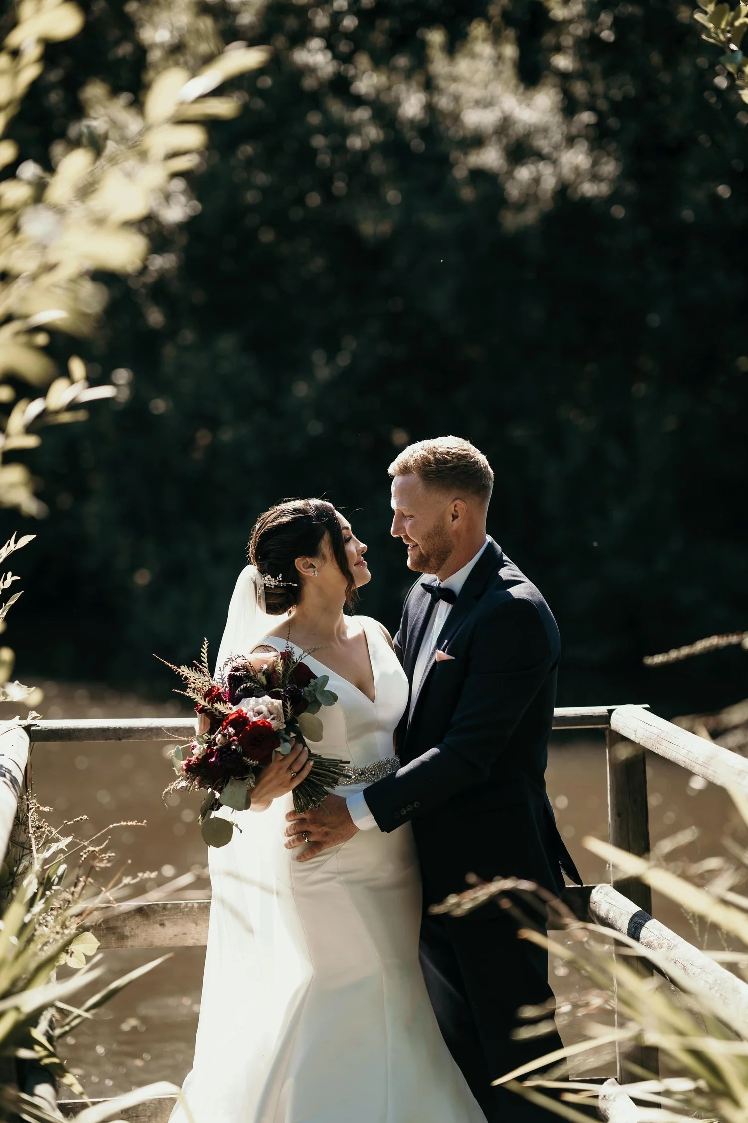 Image of the bride and groom  at the pond at Llanerch Vineyard. The bride is holding her bouquet of red burgundy and blush pink  roses l