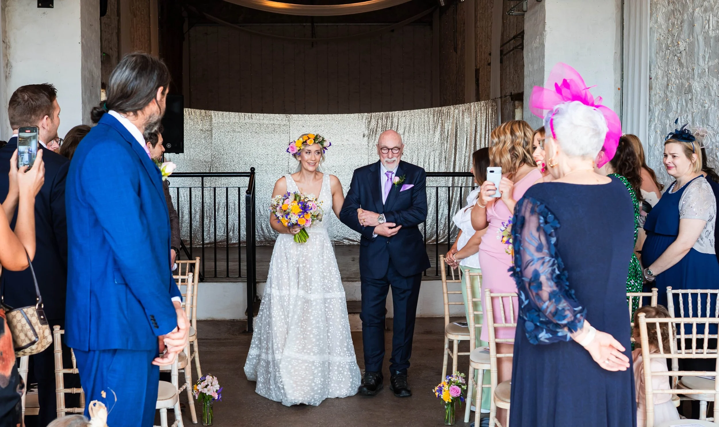 The bride is walking down the aisle at Rosedew holding her dad's arm, she is holding a colourful seasonal spring flower bridal bouquet and wearing a wedding flower crown. The aisle is dotted with jam jars of spring flowers , iris, daffodils, narcissi
