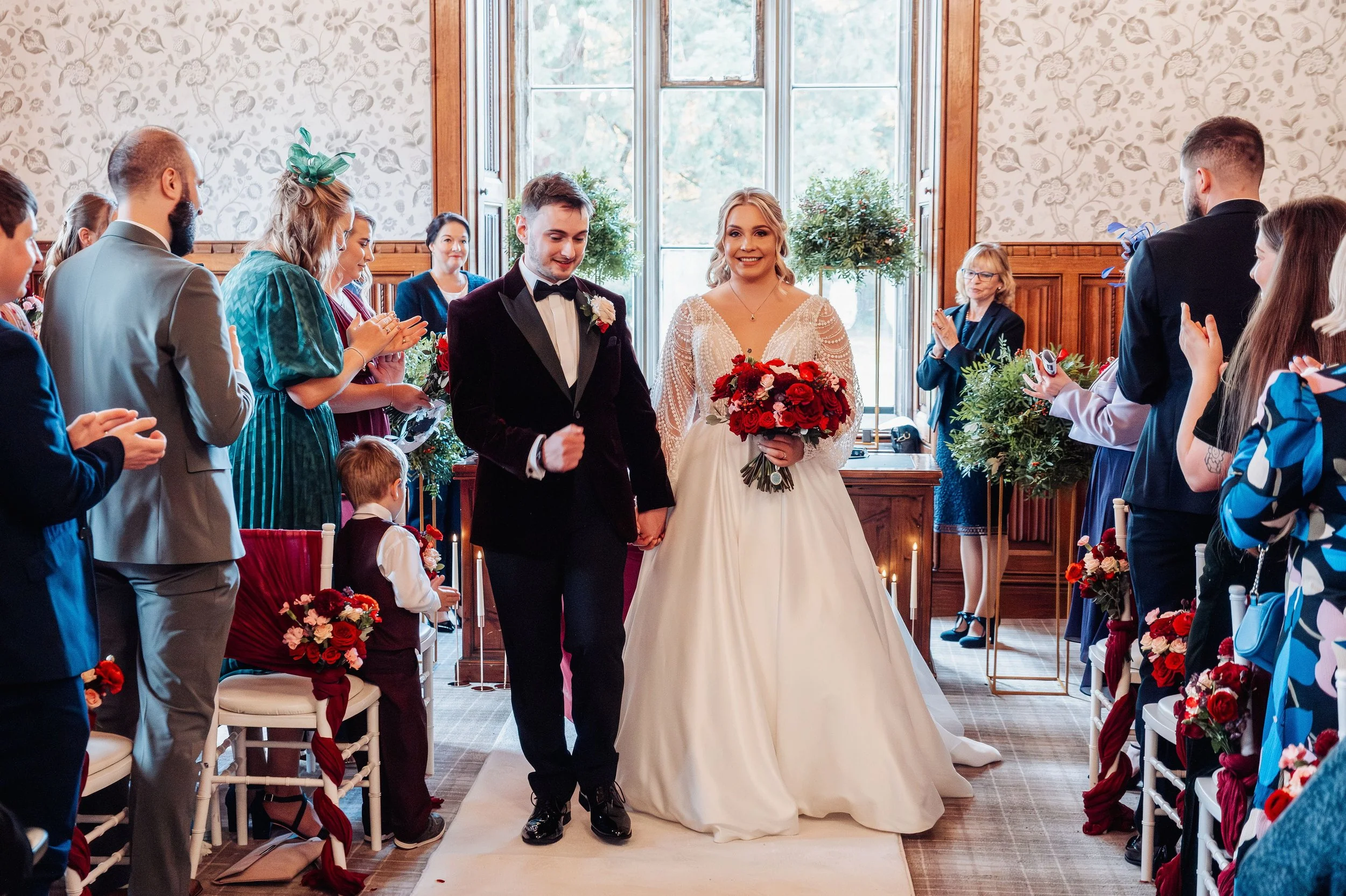 The wedded couple are walking out of the ceremony room smiling after getting married. The aisle chairs are decorated with bouquets of flowers, the foliage domes on tall gold stands are in the background.