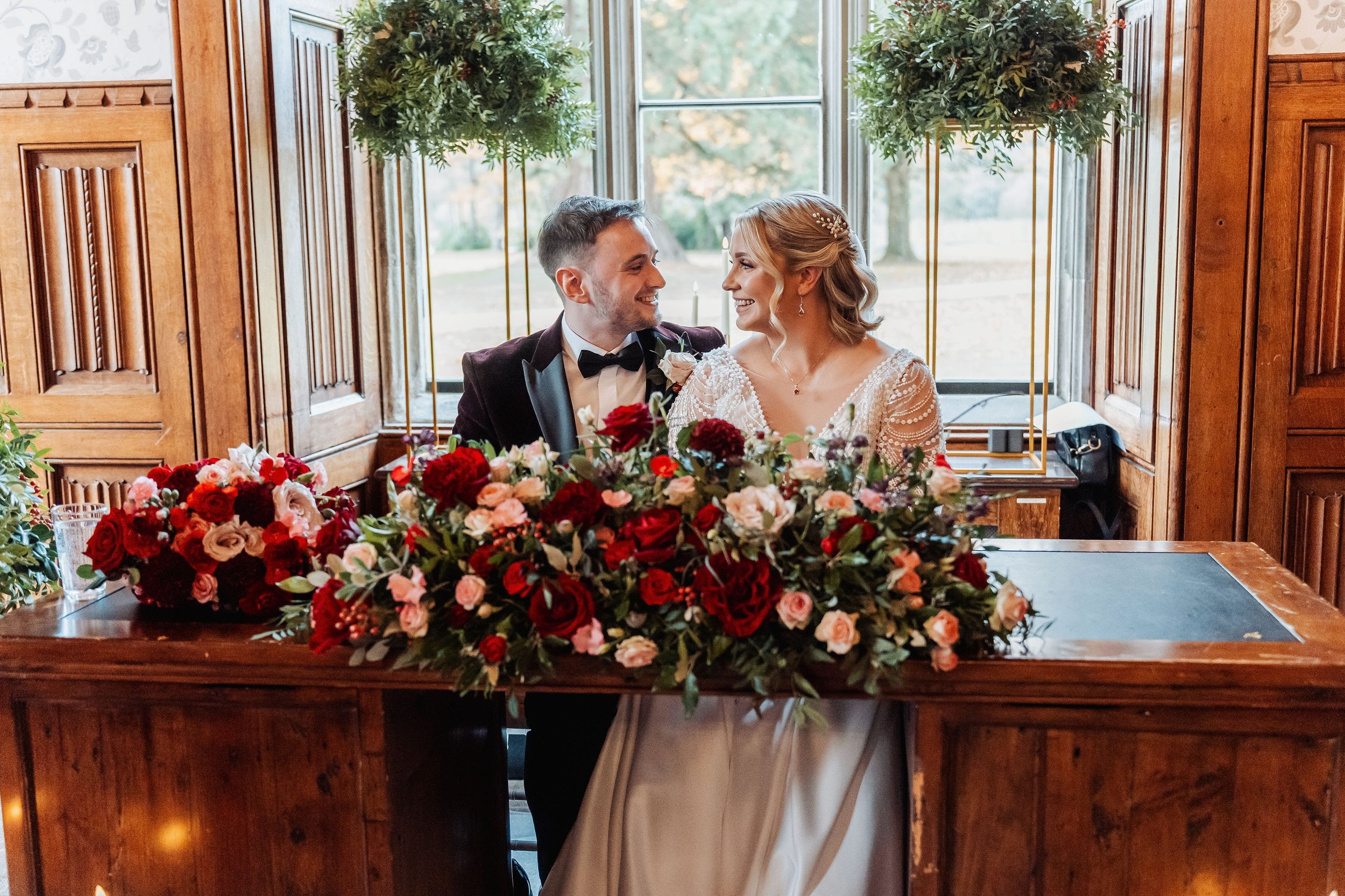 The bride and groom are sat behind the low floral arrangement of deep burgundy, reds and pinks signing the wedding register
