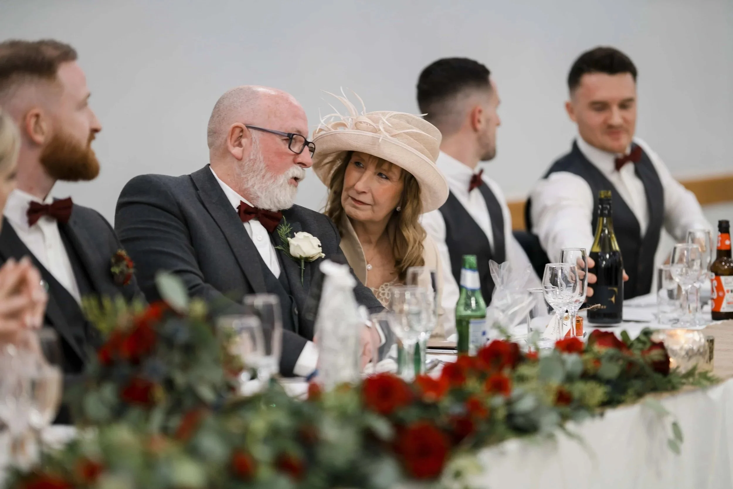 The guests are sat at the top table during the speeches. In the fore front of the picture is a garland of red roses, blue thistle and winter foliage
