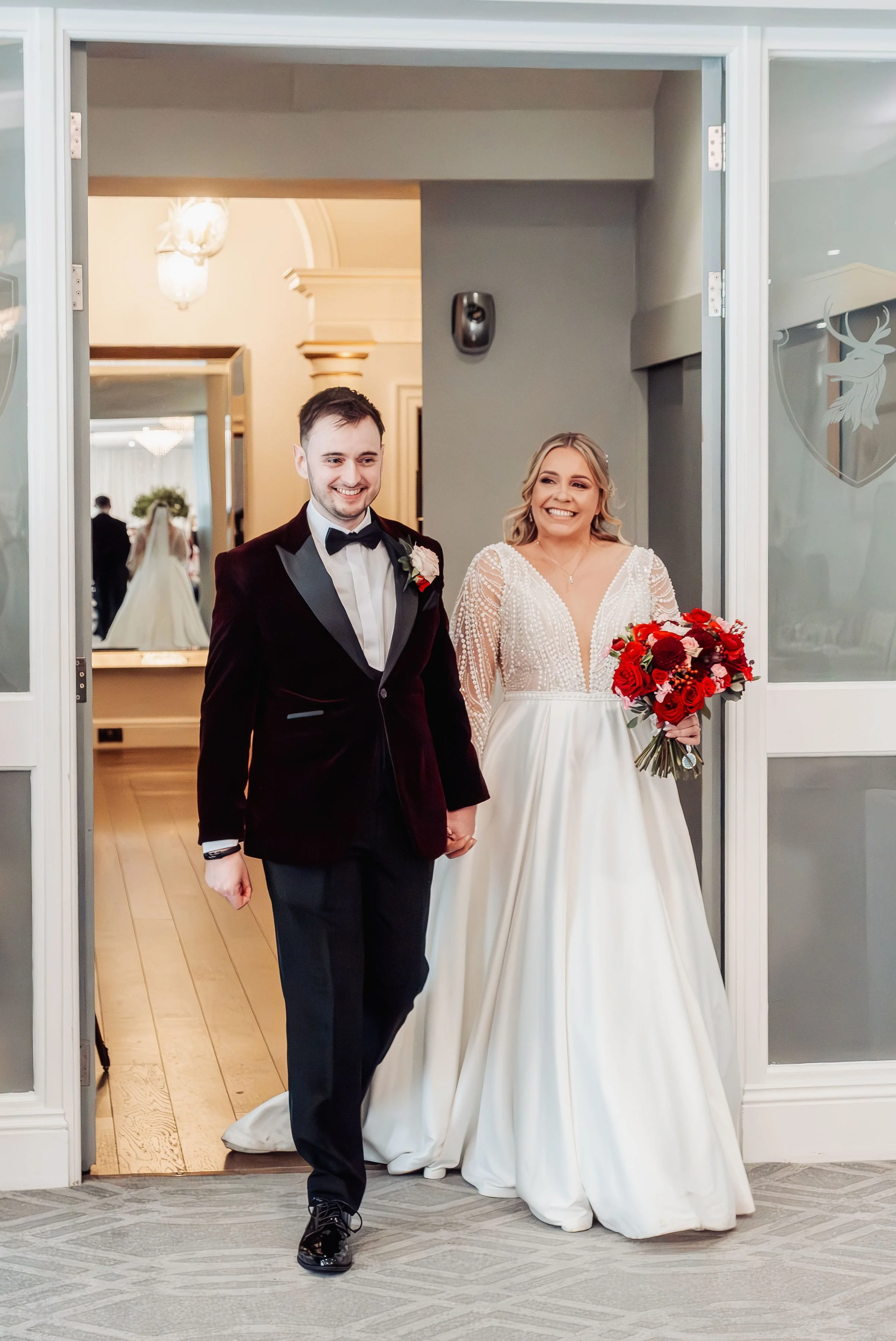 Walking into the wedding breakfast at Hensol Castle, the couple are smiling broadly, the bride is holding her bridal flowers with burgundy, red and pink flowers