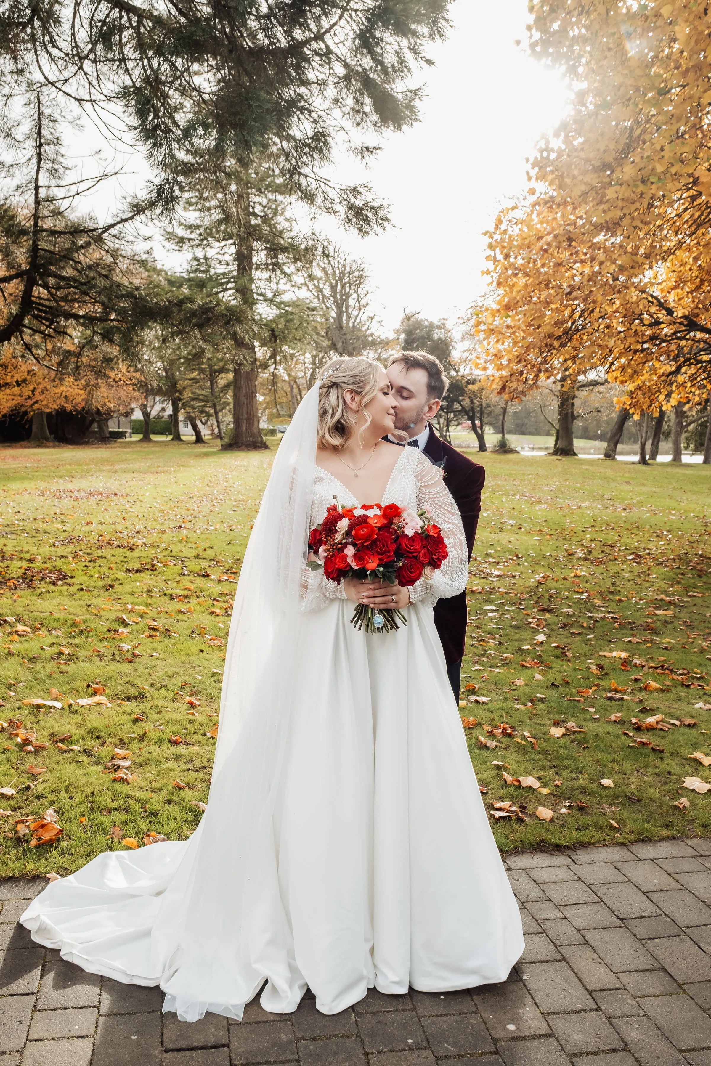 The couple are stood in the November sunshine  at Hensol Castle, the bride is holding her bouquet full of burgundy, crimson and soft pink flowers