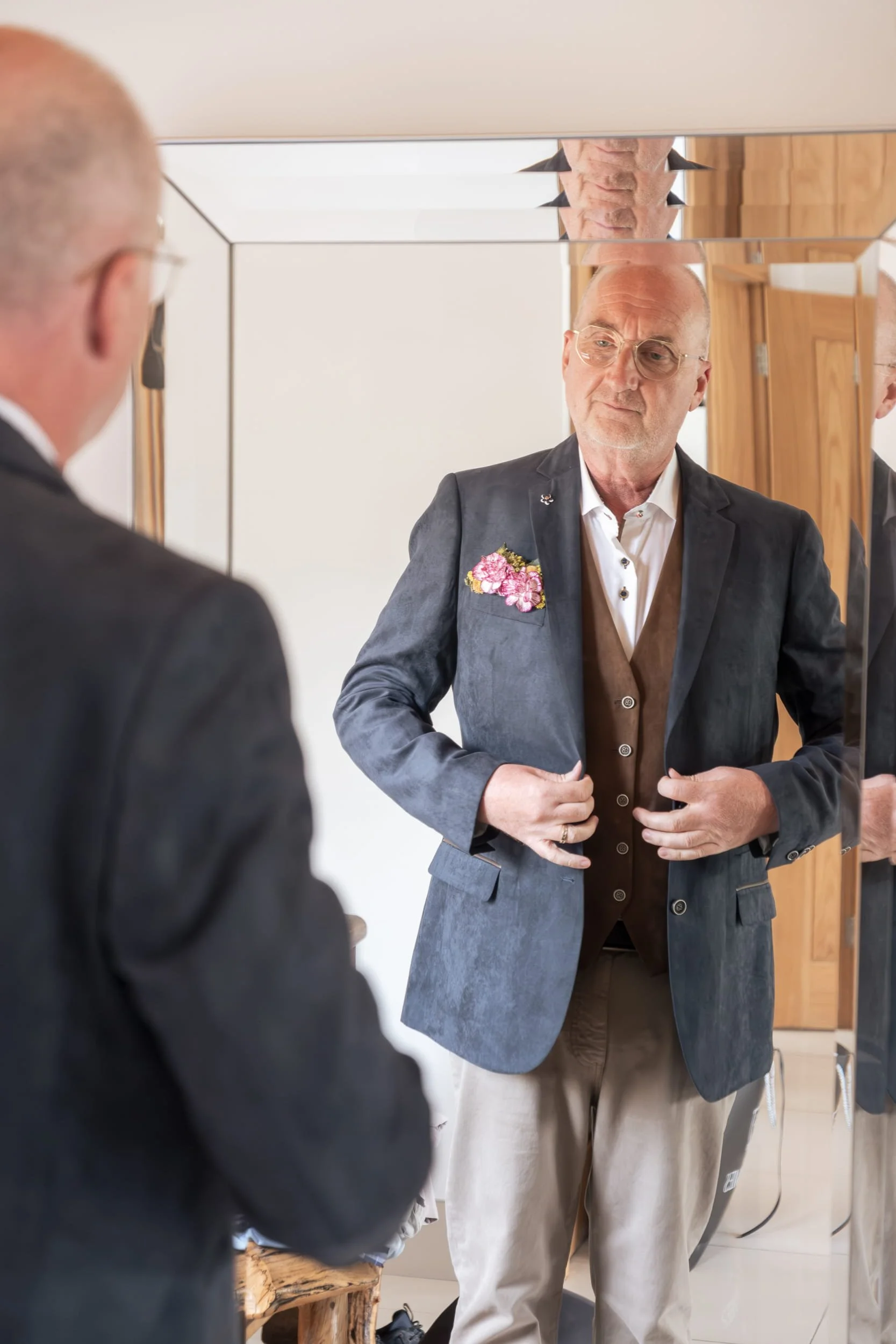 The groom is looking in the mirror at his floral pocket square with purple flower heads