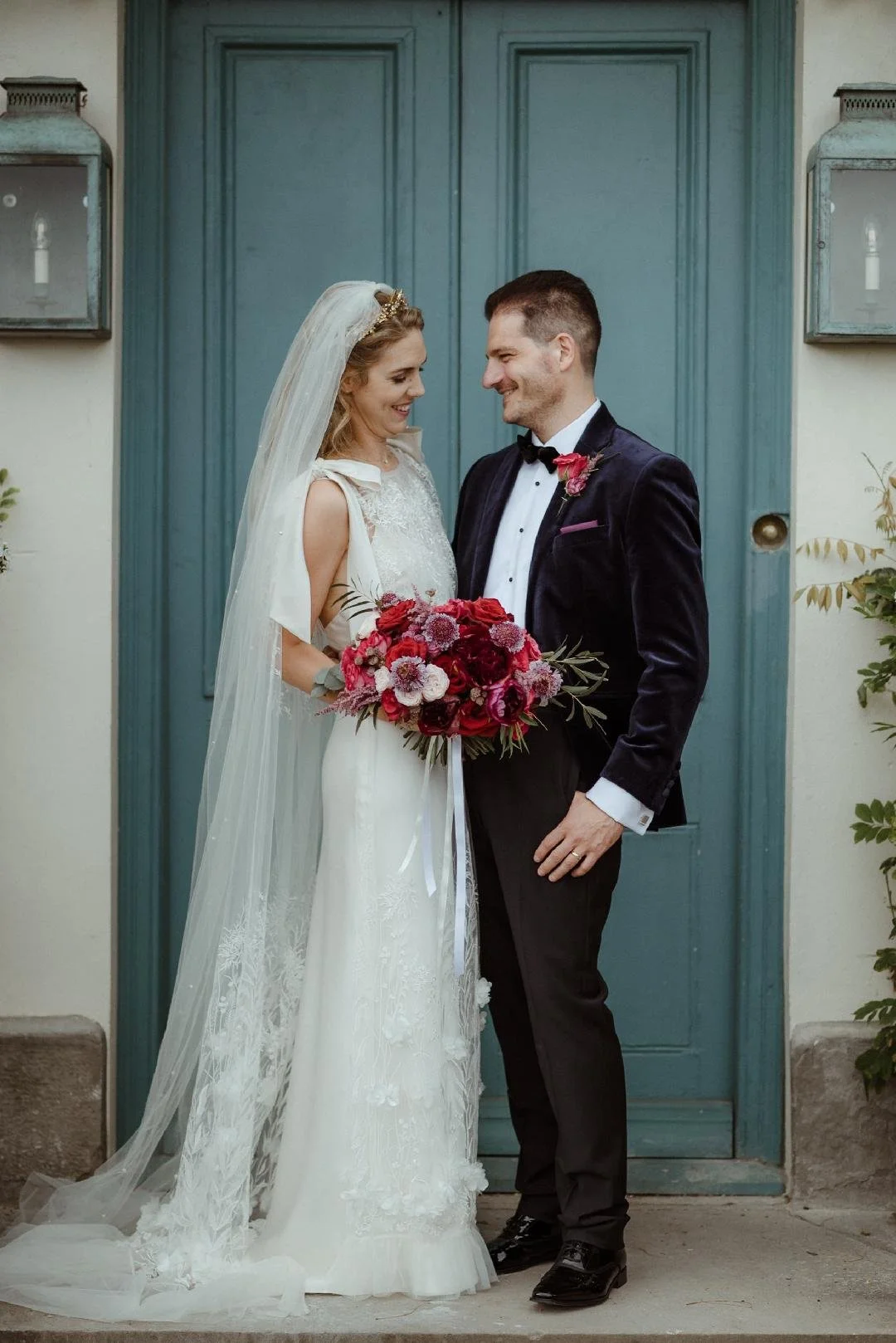 The newly weds are stood close together in front of a beautiful green/blue door, the bride is holding her bridal bouquet in burgundy, deep reds and pinks the groom wearing a matching buttonhole