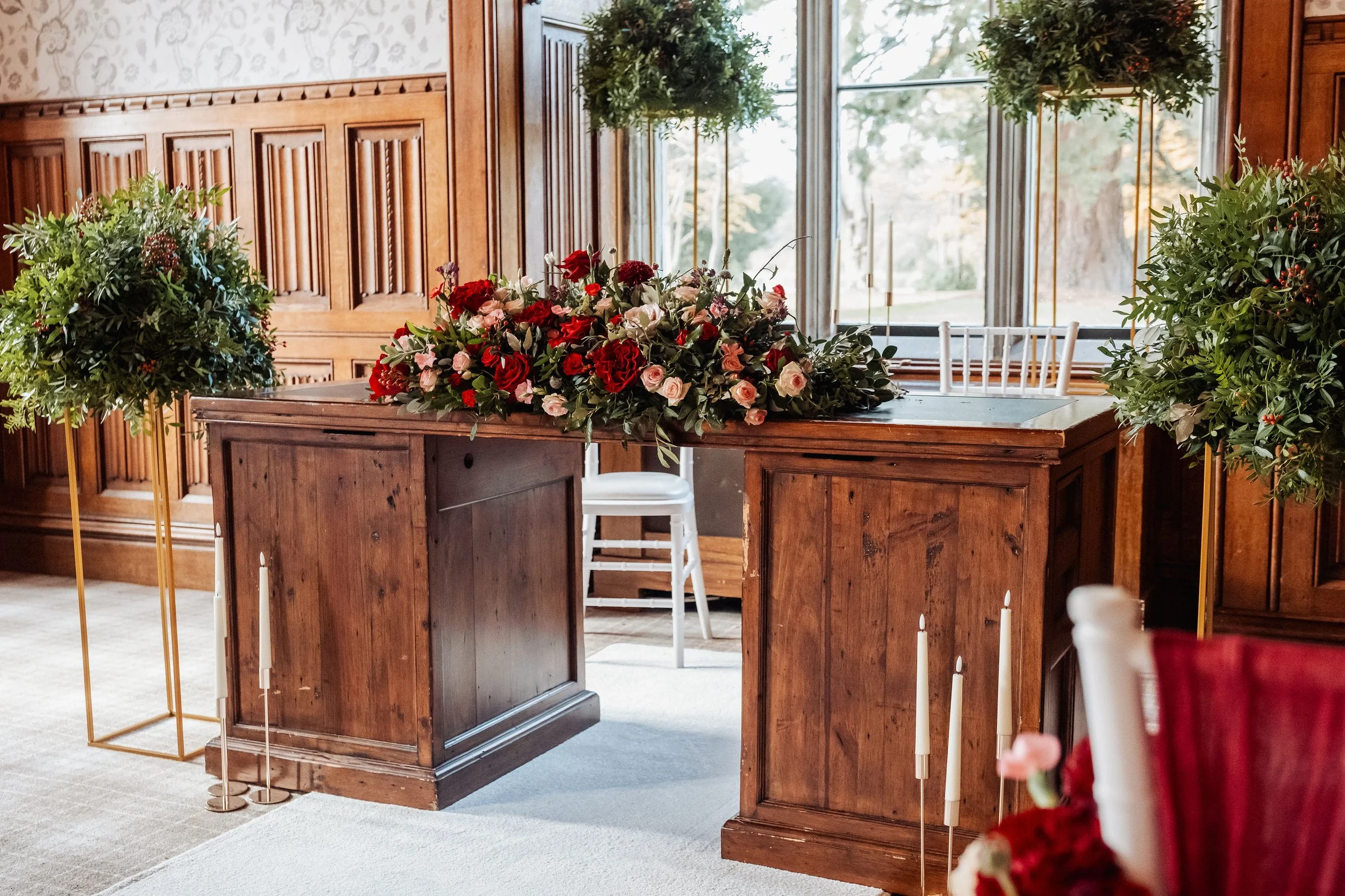 The ceremony room at Hensol Castle, tall arrangements with mounds of foliage sit beside the registry table which is adorned with a low arrangement full of seasonal flowers in colours burgundy, reds and pinks