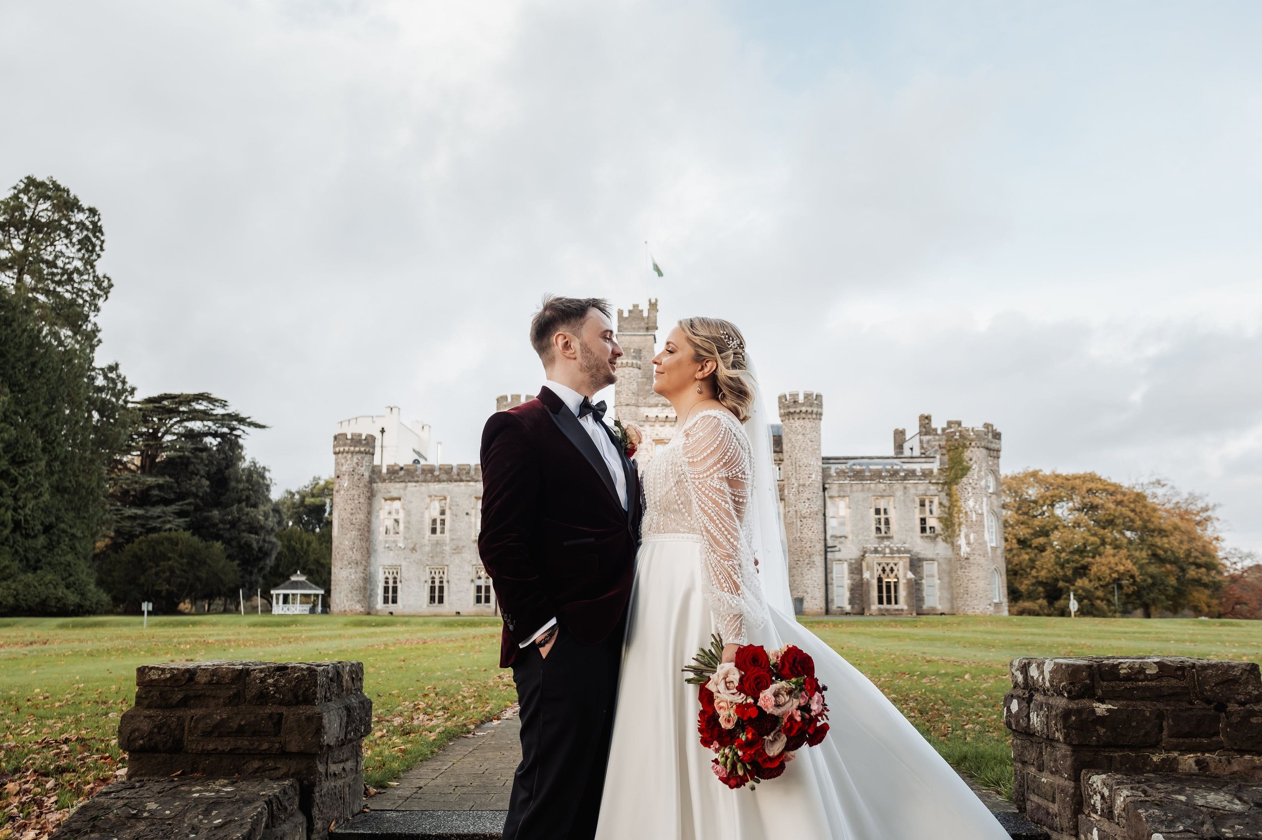 The newly weds are stood looking at each, the bride is holding her bouquet full of burgundy, crimson and soft pink flowers, Hensol Castle is in the background