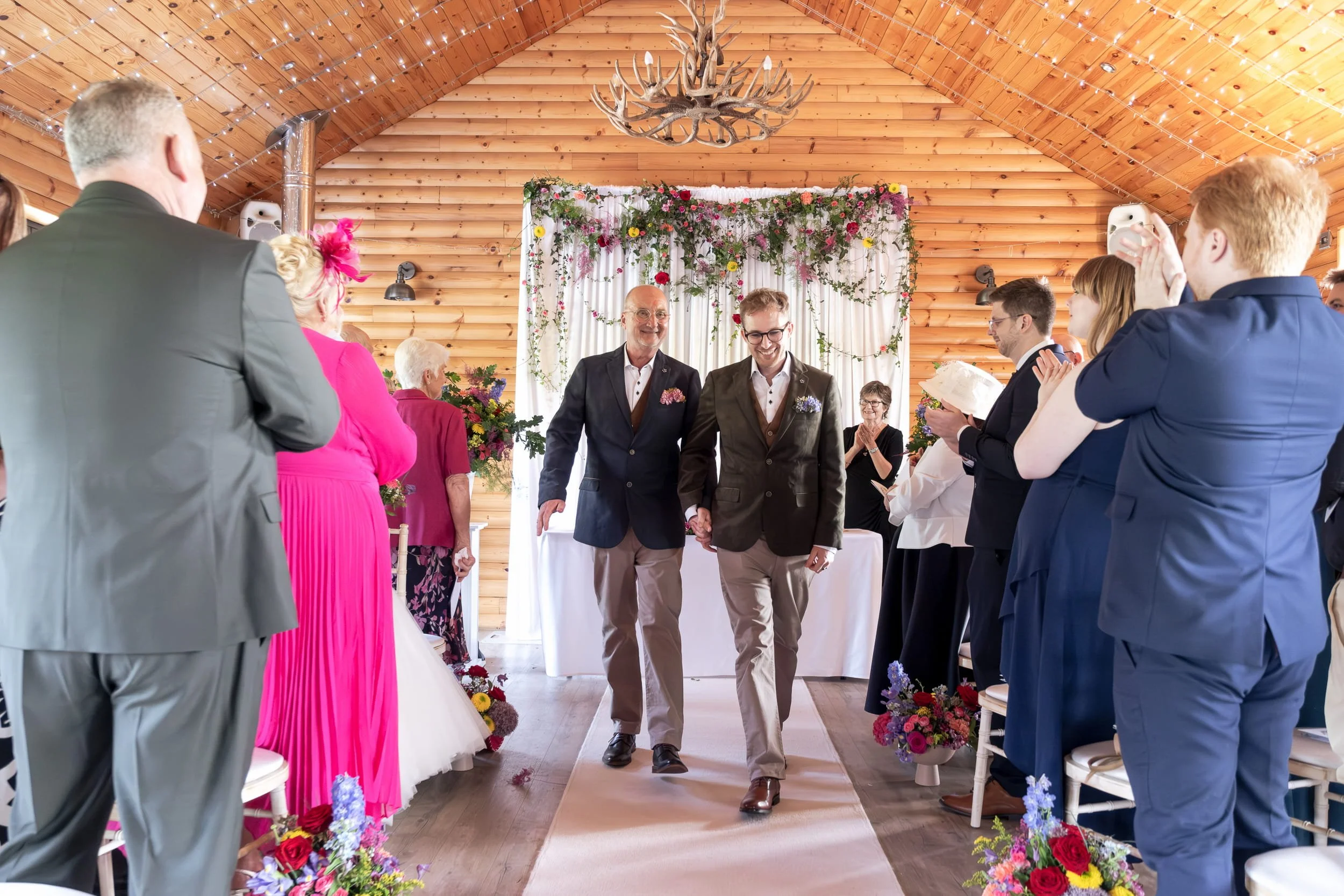 Just married gay couple walking down the aisle in front of floral backdrop at Rhodri's by the Lake