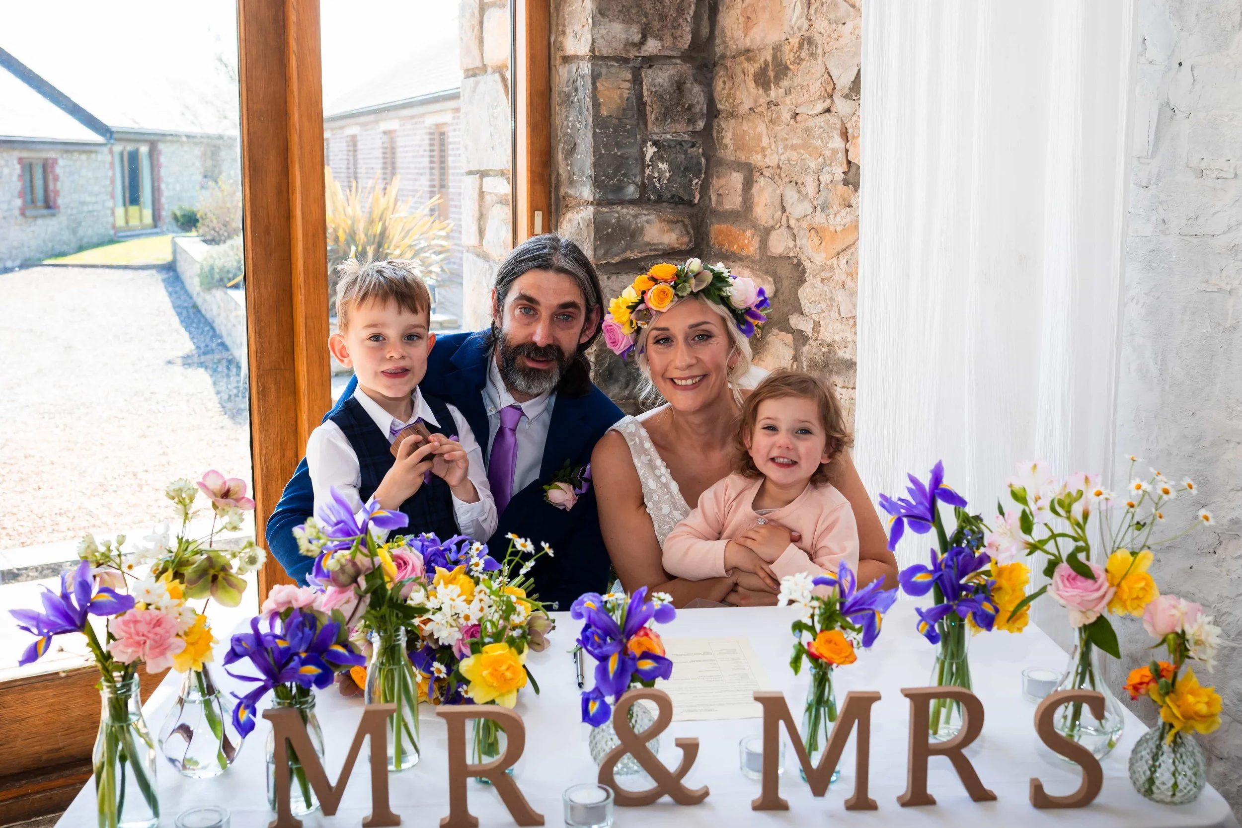 The just married family at Rosedew farm sat at a table full of mix and match bud vases full of spring flowers , iris, daffodils, daisies, narcissi, helebores, roses