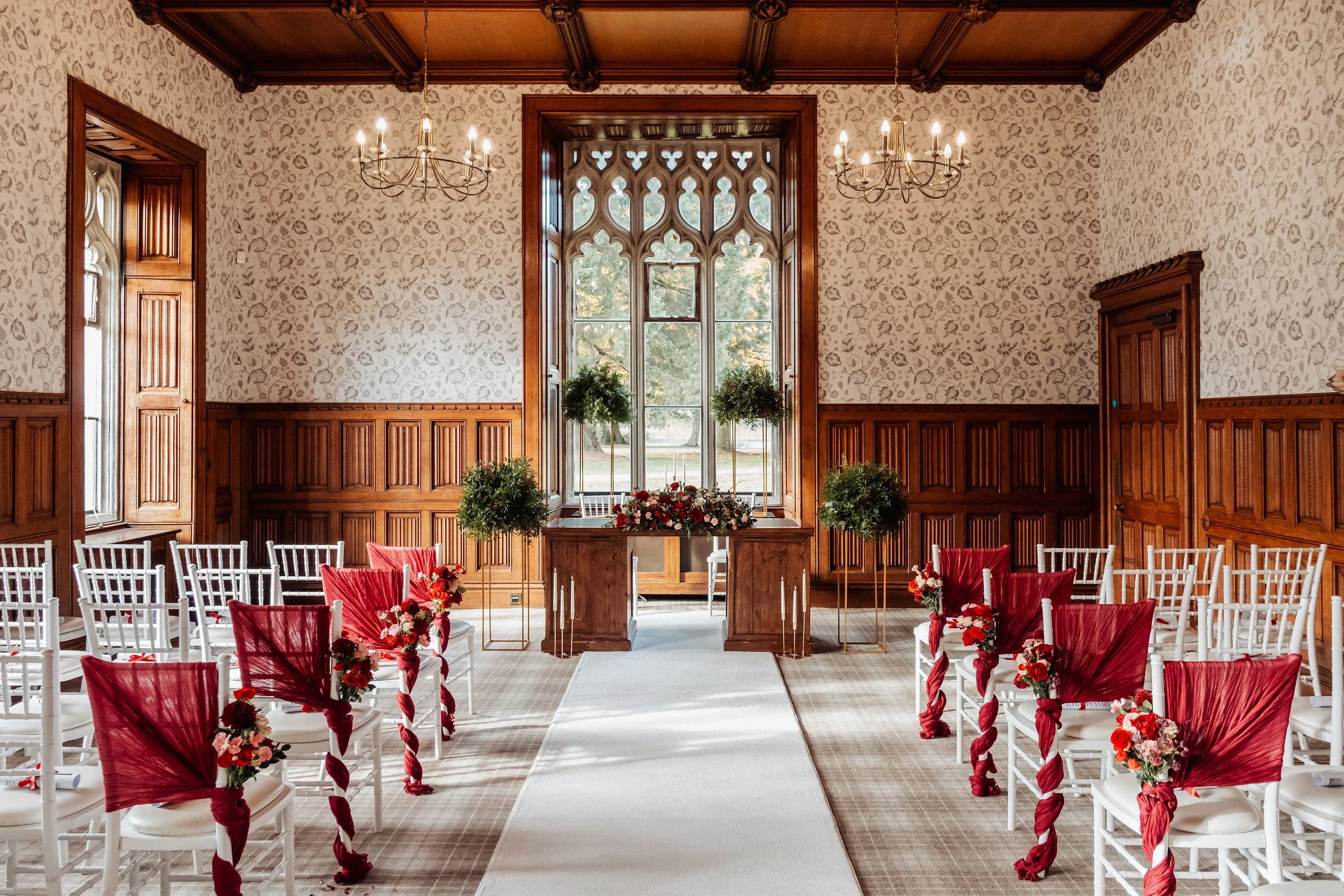 The ceremony room at Hensol Castle, floral bouquets in burgundy, reds and pinks are tied to the chairs, tall arrangements with mounds of foliage sit beside the registry table which is adorned with a low arrangement full of seasonal flowers
