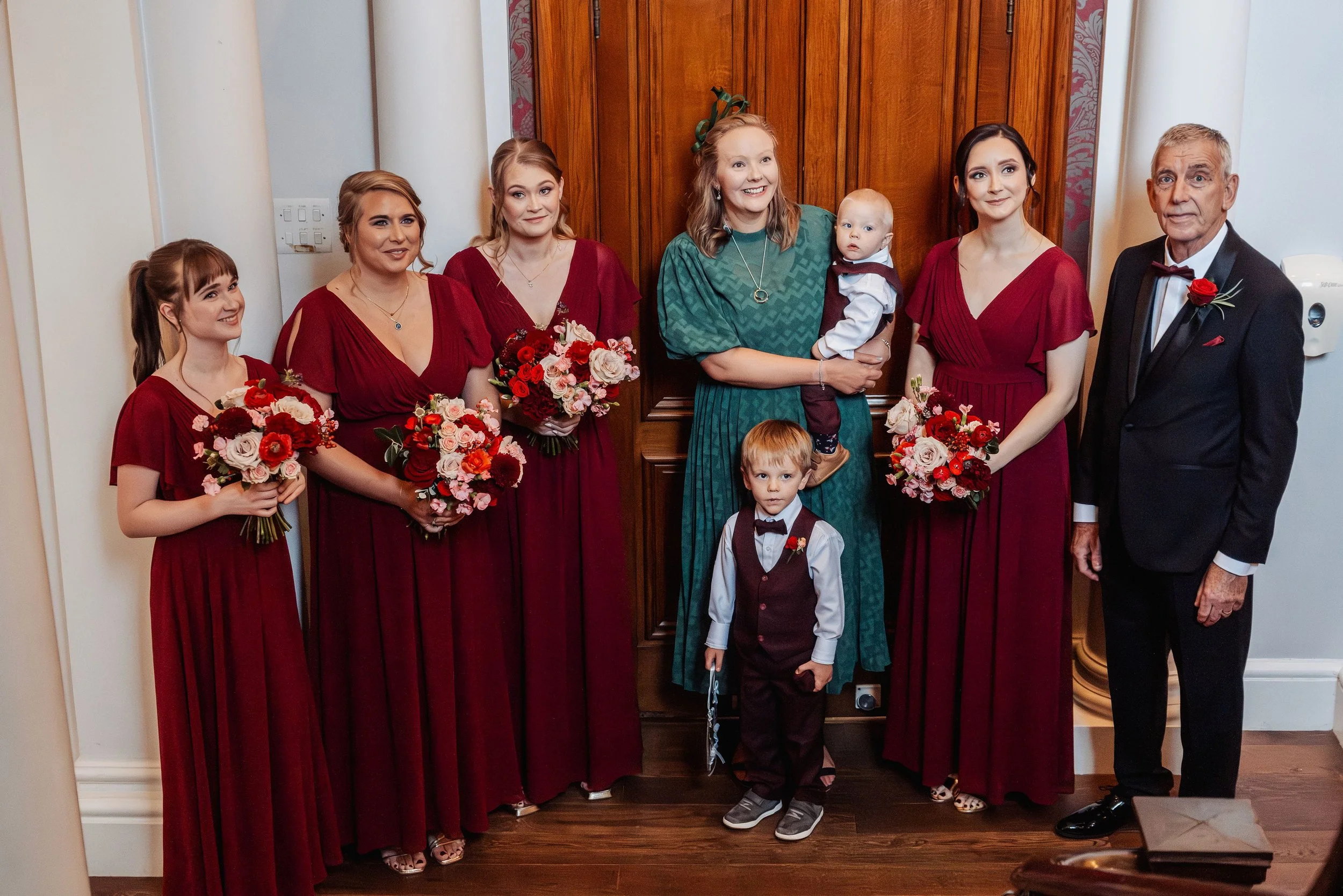 The bridesmaids are stood at the bottom of the stairs waiting for the bride reveal holding their bouquets in burgundy, crimson and soft pinks