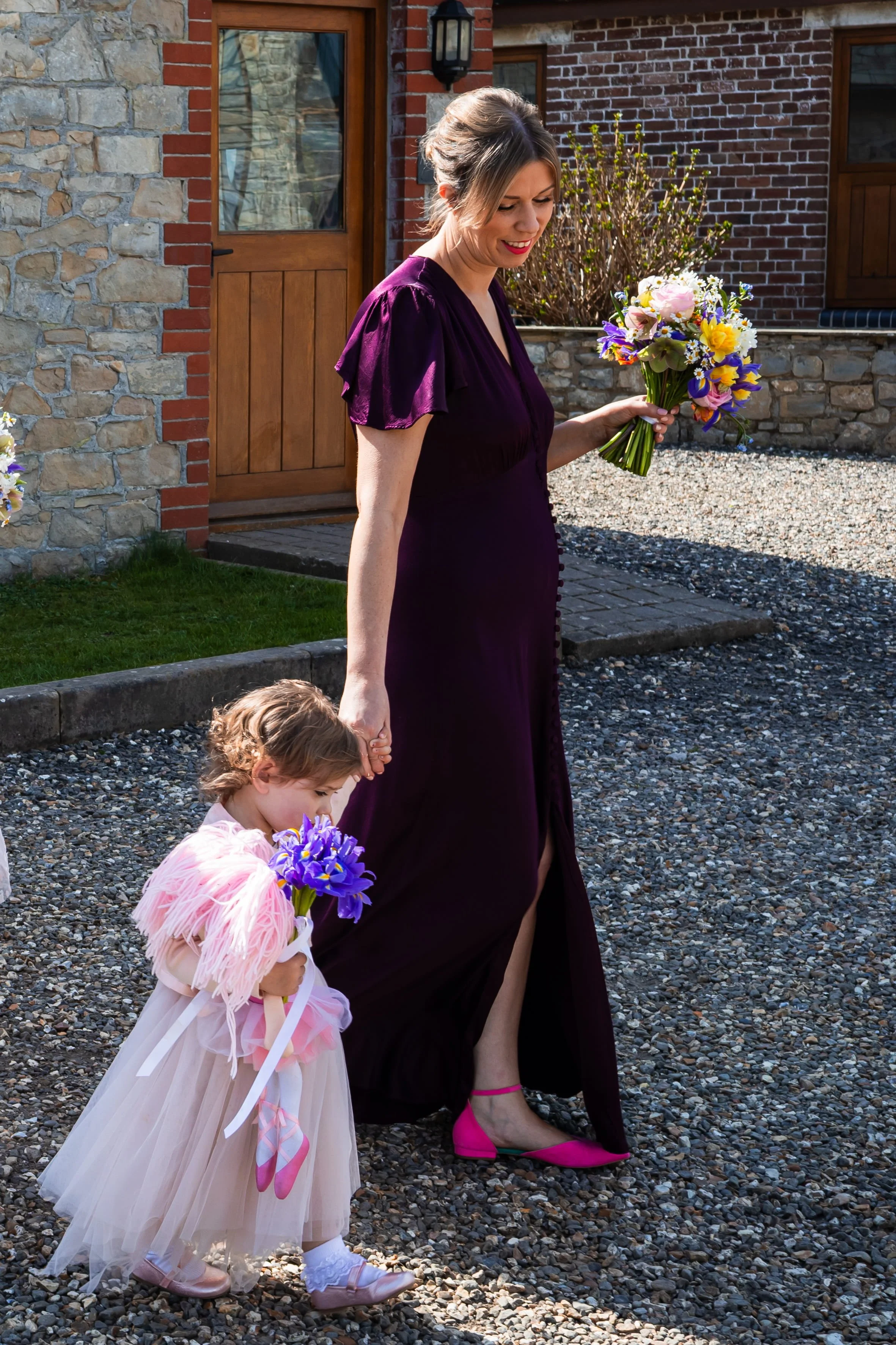The flower girl is carrying a posy of iris flowers and holding the hand of a bridesmaids who is carrying her bouquet full of colourful, seasonal spring flowers - daffodils, iris, forget me not, helebores, narcissi