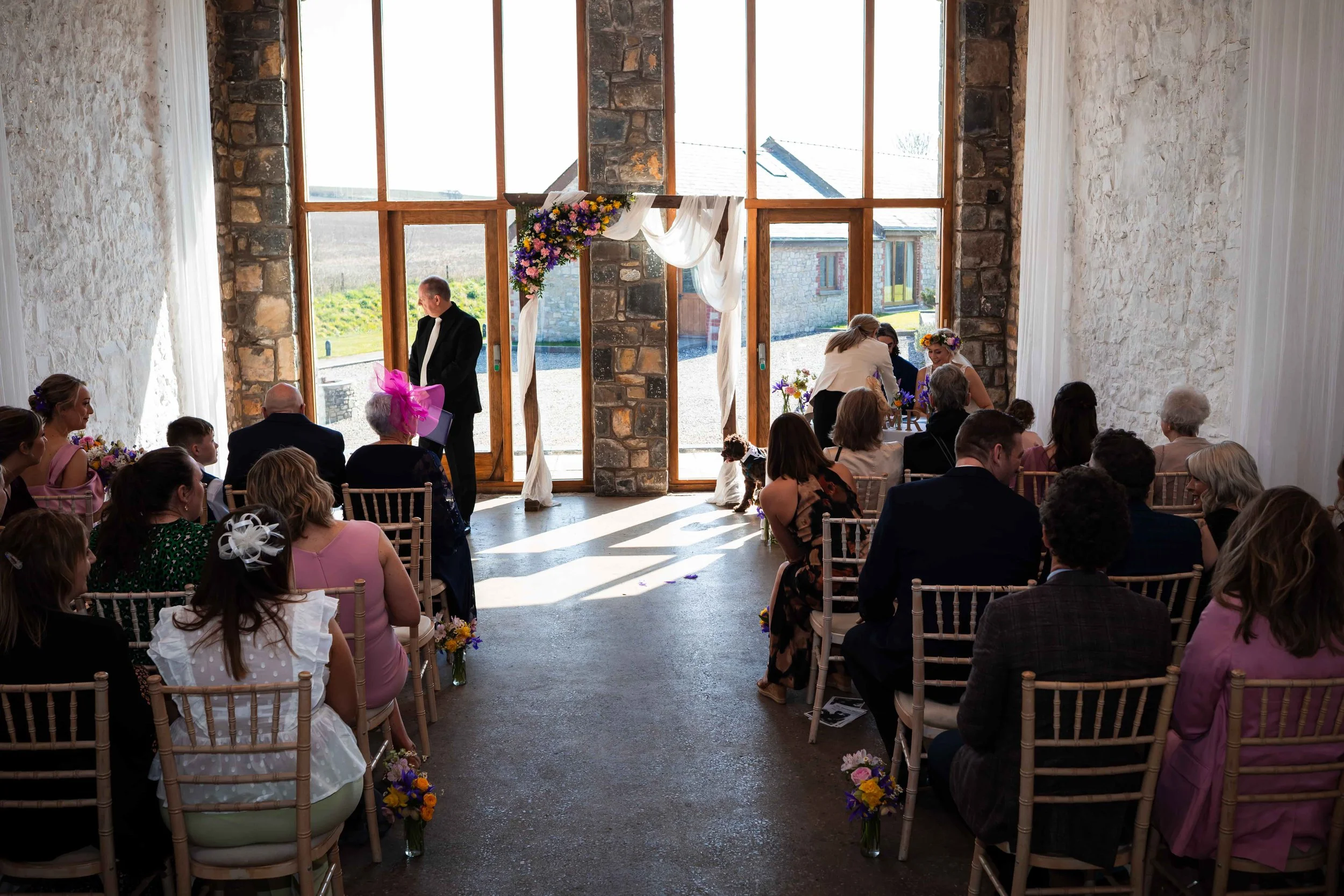 The ceremony room set up at Rosedew Farm with the arch garland of seasonal flowers of colourful spring iris, narcissus, astrantia, daisies, helebores, daffodils and roses. The floor is lined with small jars full of spring wedding flowers