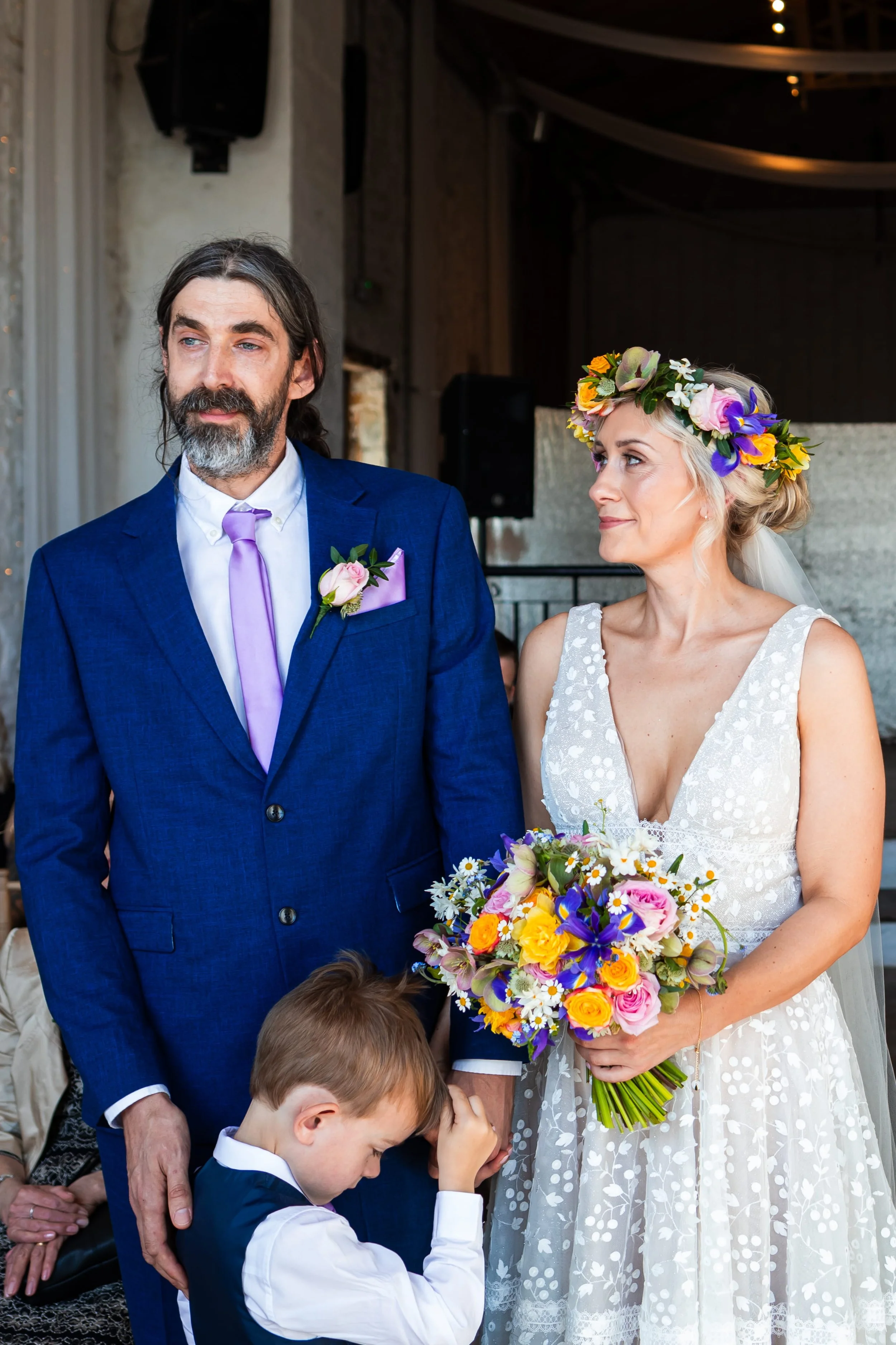 The bride and groom are stood saying their vows at Rosedew farm. The bride looks stunning wearing her full bridal floral crown of colourful, seasonal spring flowers and holding her natural, spring wedding flower bridal bouquet