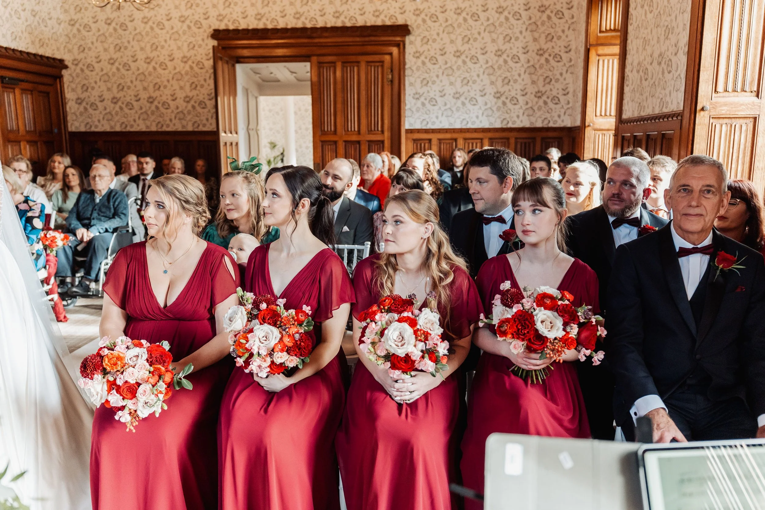 The bridesmaids are sat in the front row of Hensol Castle holding their bouquets in burgundy, crimson and pink colours watching the wedding vows