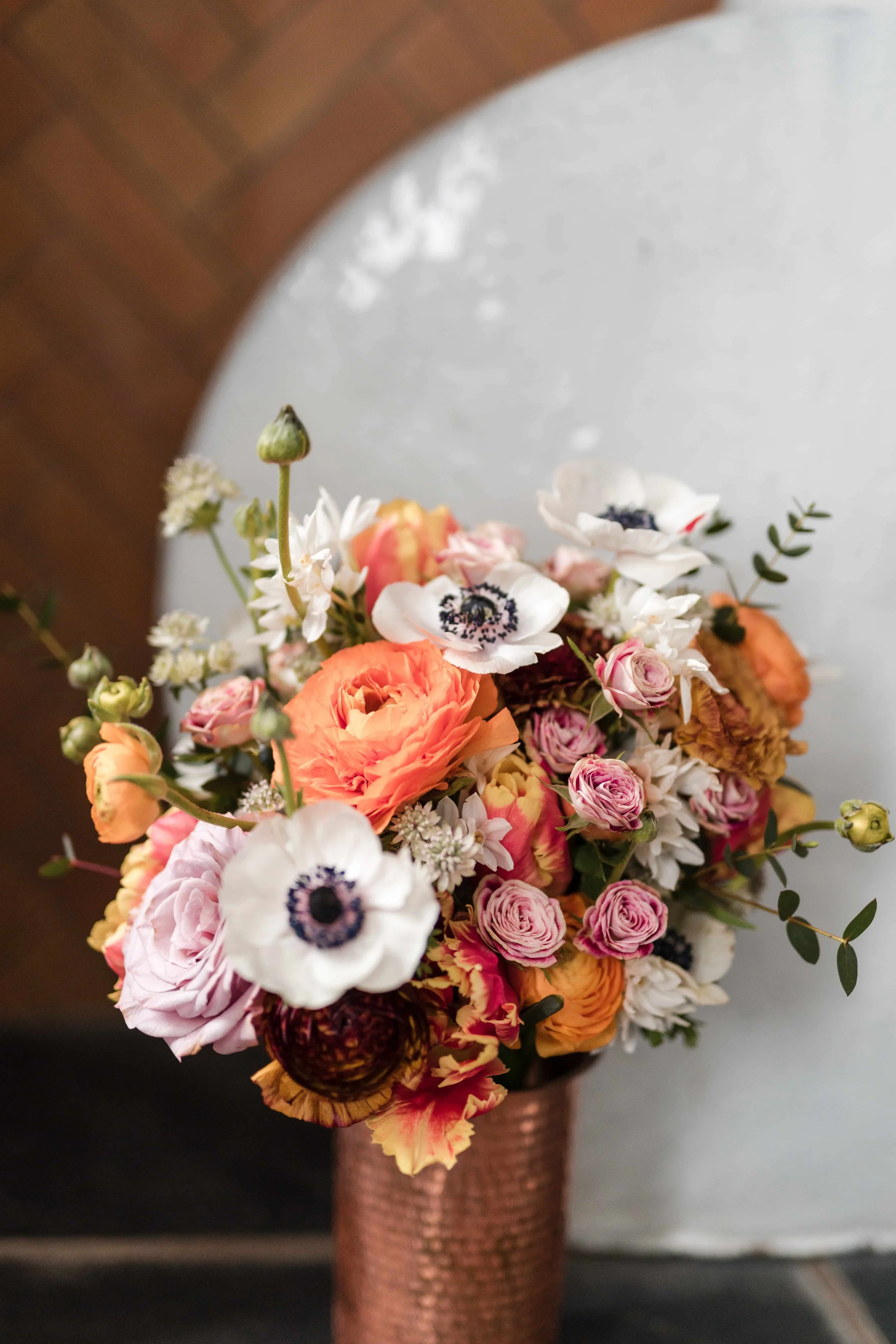 Bridal bouquet with colourful seasonal spring flowers in oranges and pinks with added white anemones sat in a copper vase