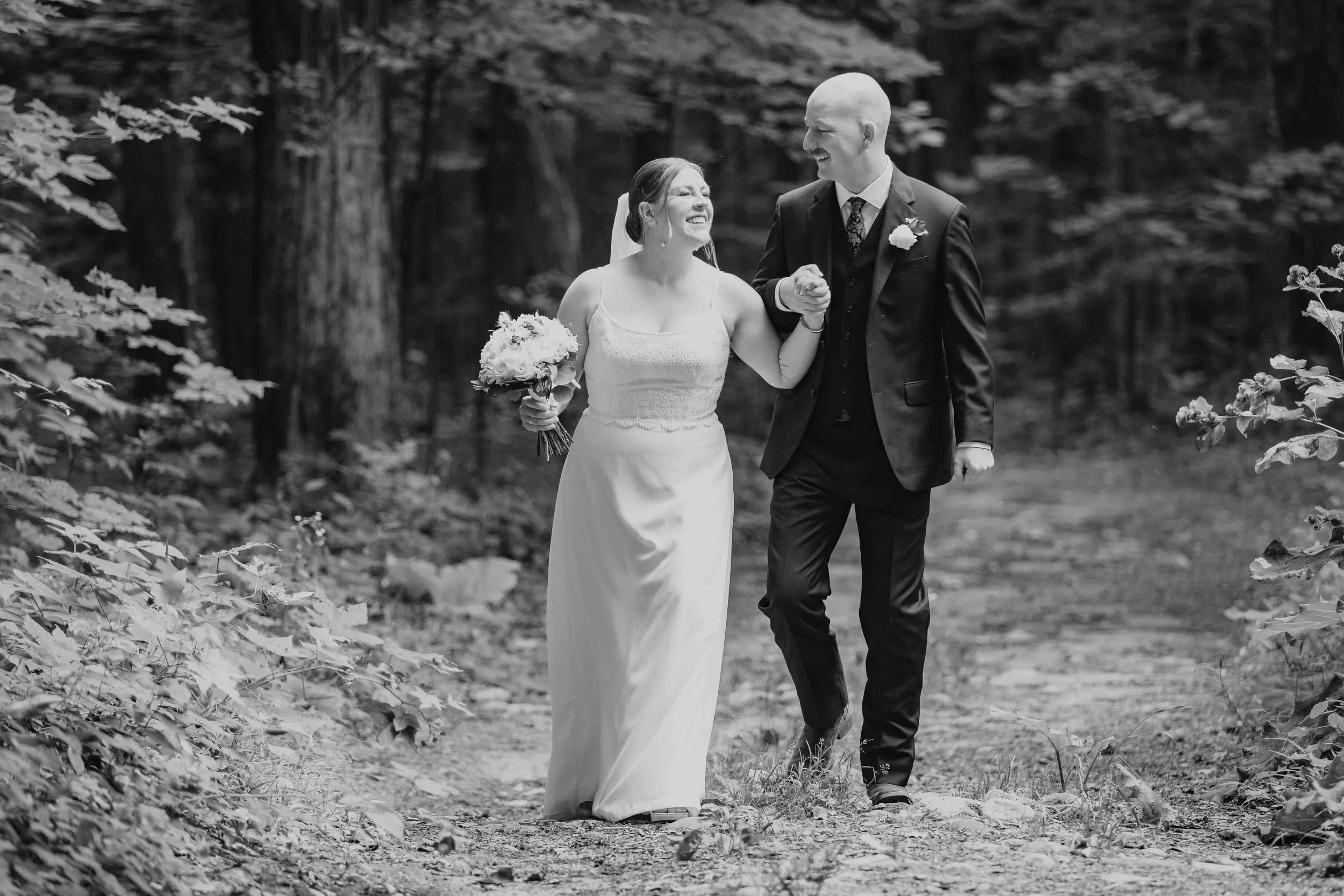 Black-and-white photo of newlyweds holding hands and laughing on a forest path at Sucrerie de la Montagne.