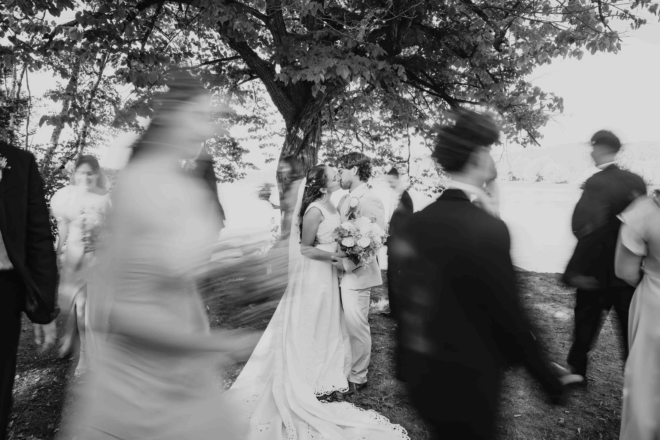 Black-and-white wedding portrait of Emma and Timothée kissing beneath a tree at Camp Livingstone, with their wedding party blurred around them