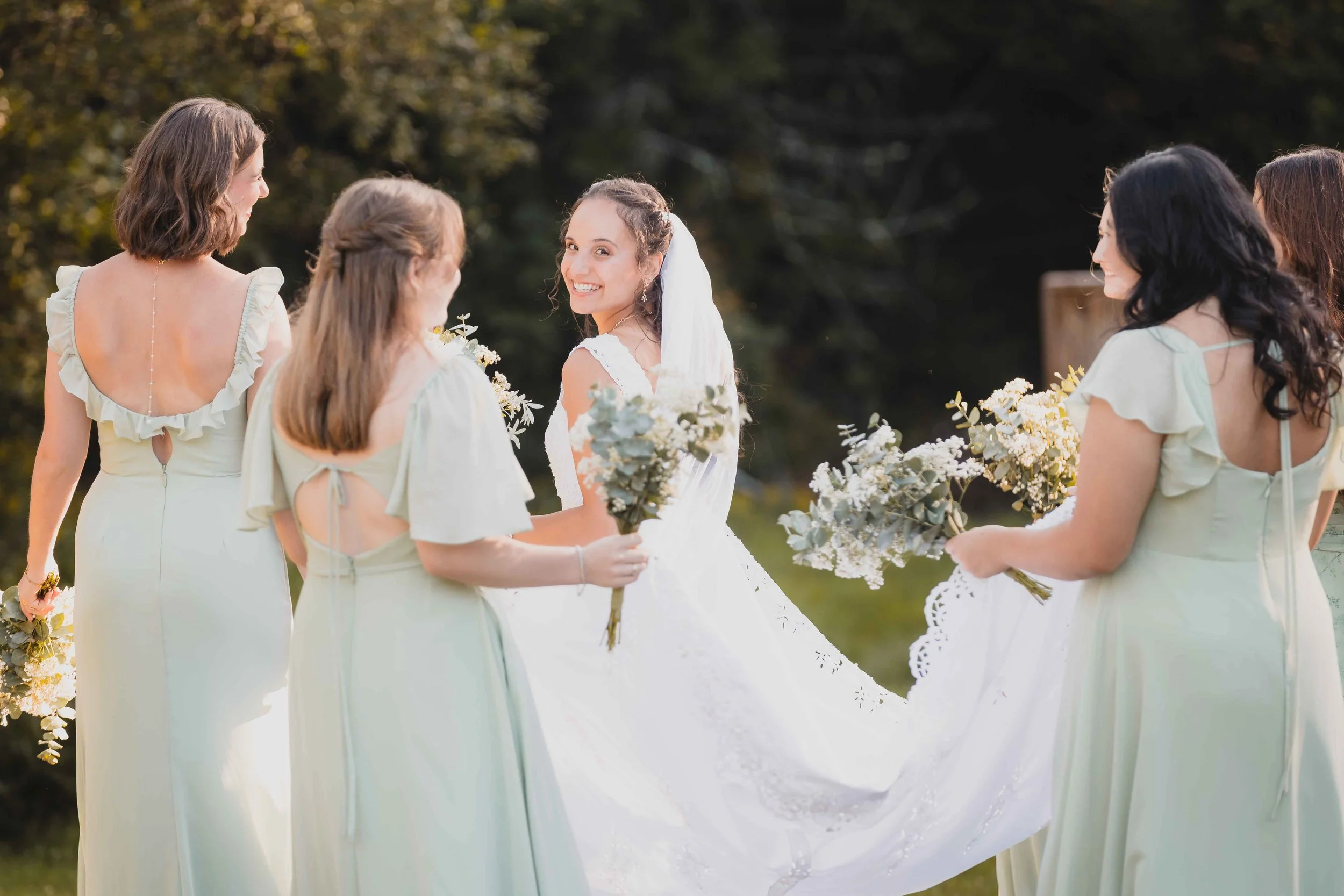 Bride smiles over her shoulder beside bridesmaids in sage-green dresses holding bouquets at Camp Livingstone in Quebec’s Eastern Townships.
