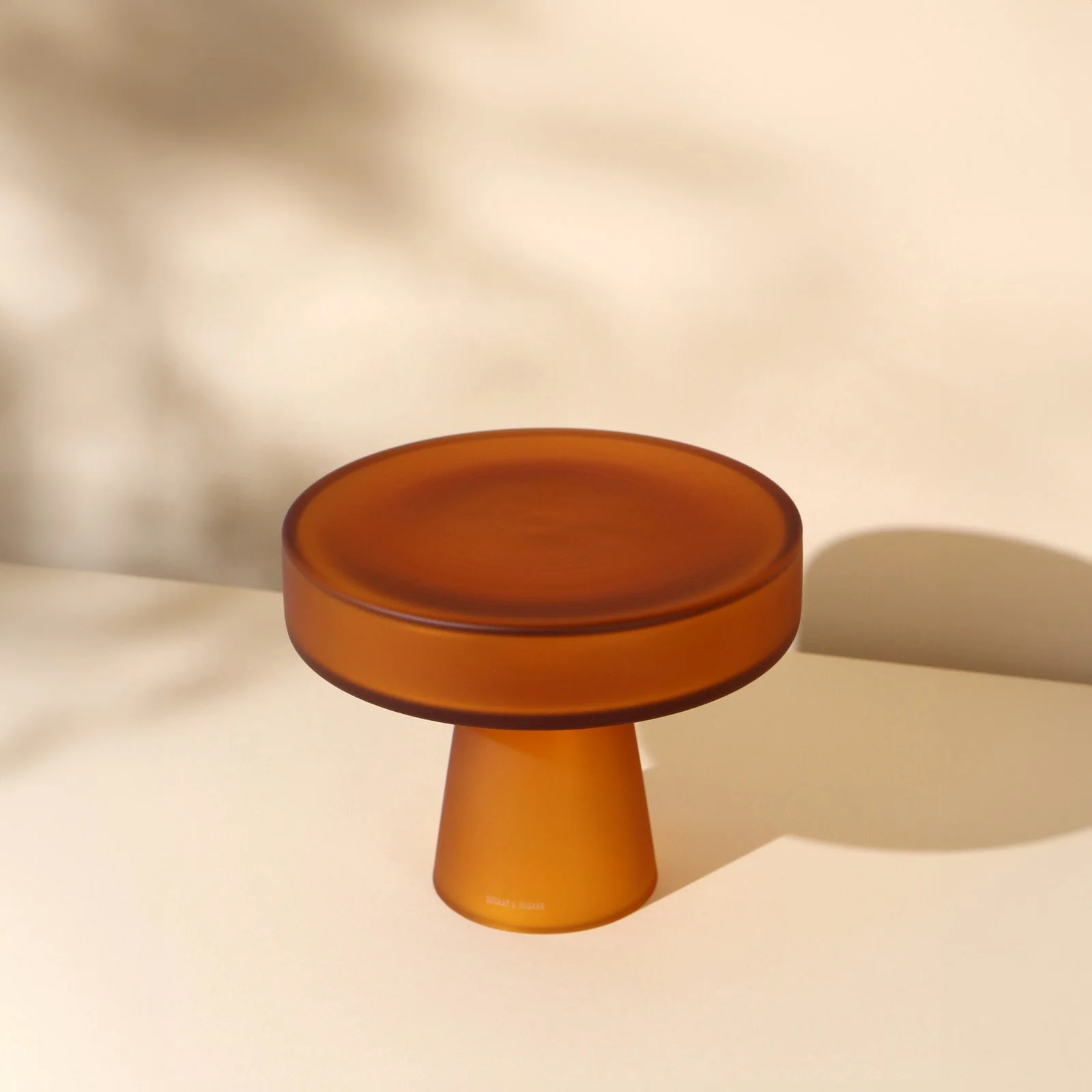 Matte amber glass cake stand on a beige background with plant shadows.