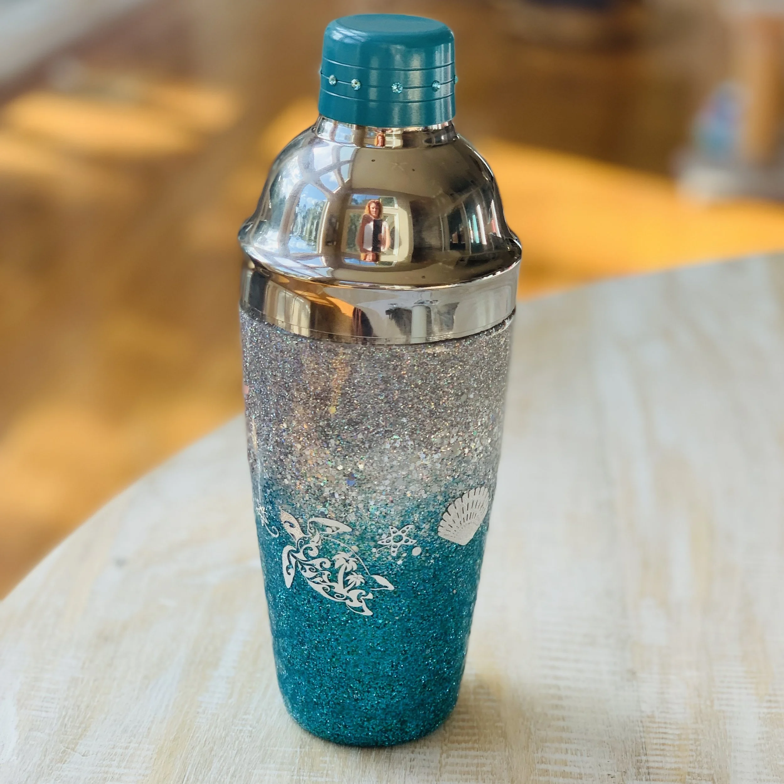 A glittery blue and silver insulated tumbler with a seashell and ocean-themed design on it, resting on a light-colored wooden surface.