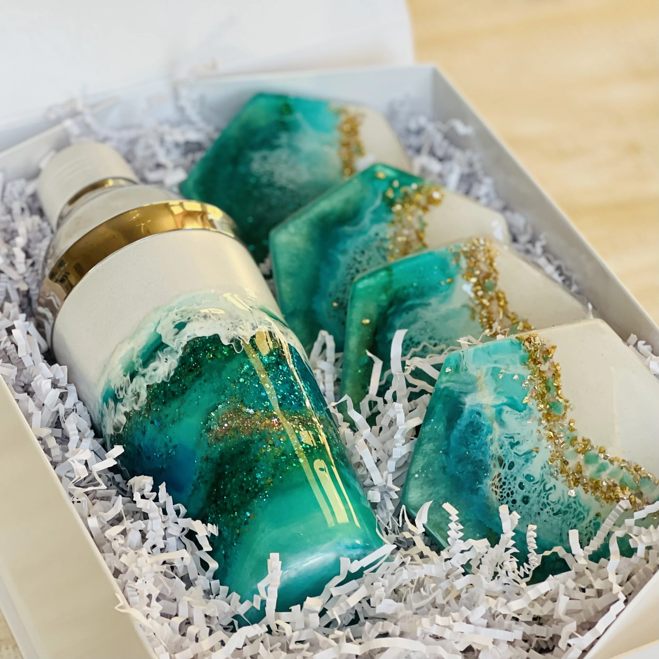A gift box containing resin art pieces with blue, teal, white, and gold accents, arranged on shredded paper.