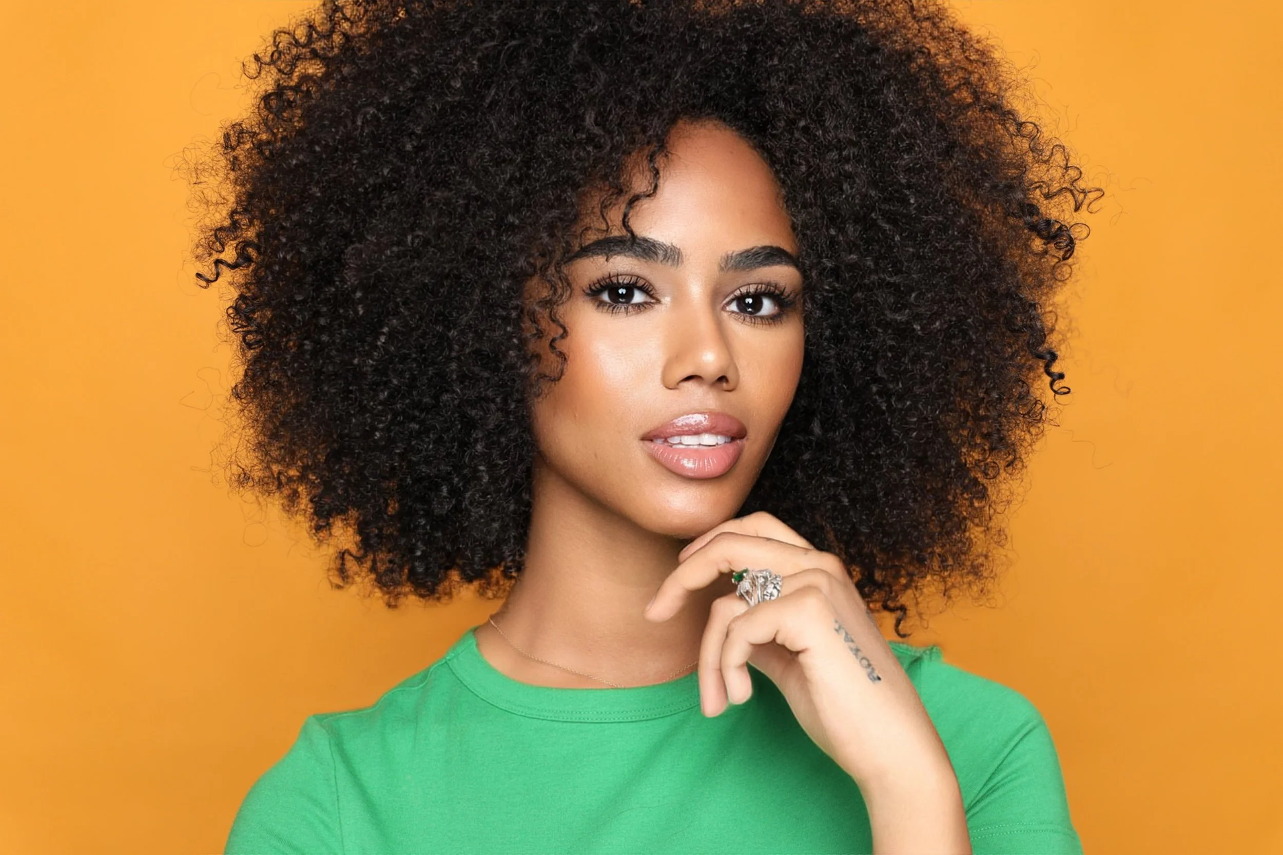 Portrait of a woman with curly hair, wearing a green top, standing against a yellow background.