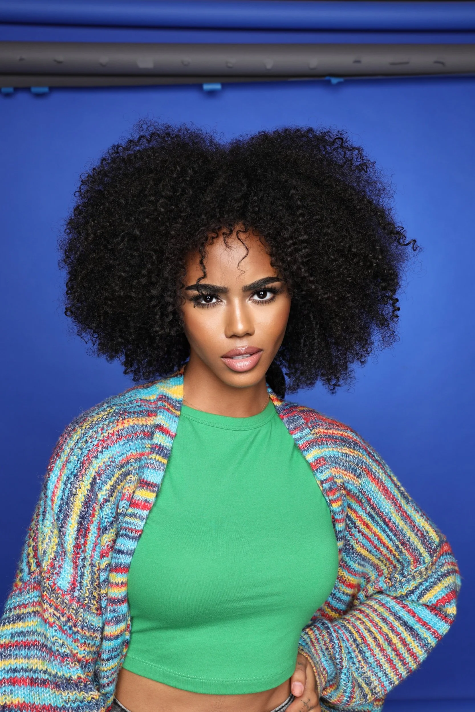 A woman with dark curly hair, wearing a vibrant rainbow cardigan over a green top, stands in front of a blue background.