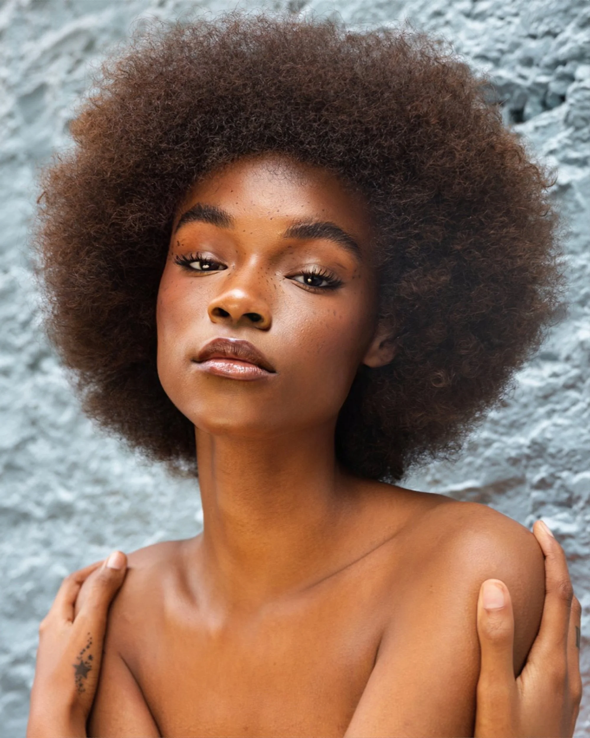 Close-up of a woman with natural curly hair posing in front of a stone or concrete background, with her hands gently touching her shoulders.