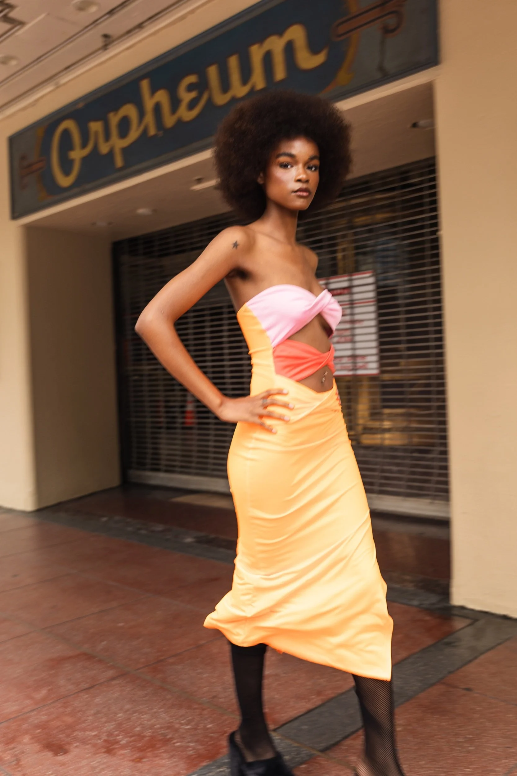 A woman with a large afro hairstyle standing outdoors in front of a closed shop with a sign that says 'Cupheury'. She is wearing a strapless color-block dress in pink, orange, and yellow, with black fishnet stockings and black shoes. She has a confid