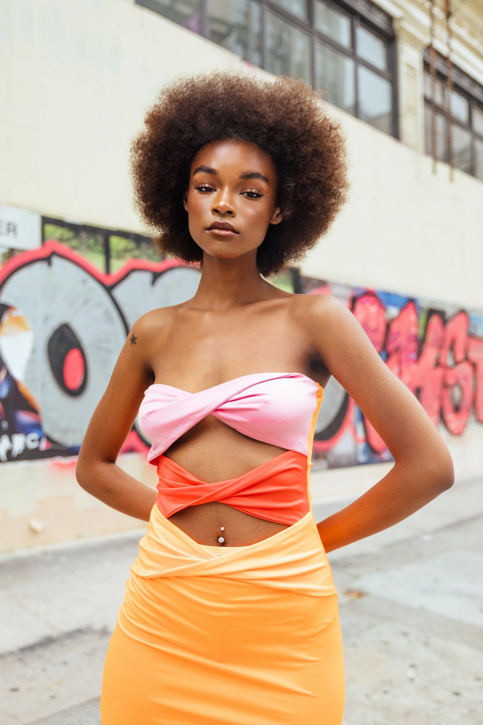 A young woman with dark skin and an afro hairstyle standing in front of a graffiti-covered wall, wearing a colorful strapless top and a matching skirt.