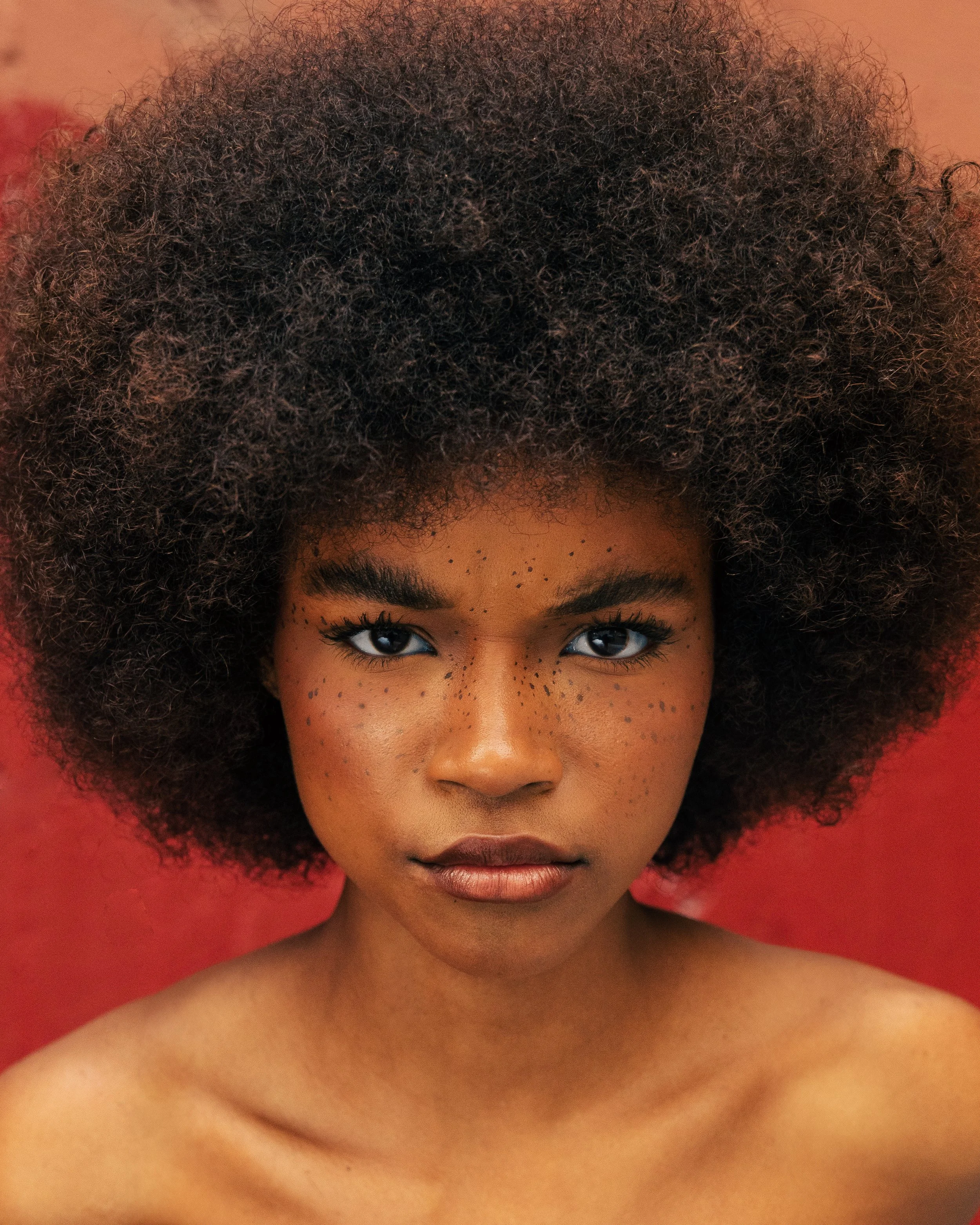 Close-up of a woman with dark skin, freckled face, and curly afro hair, against a red background.