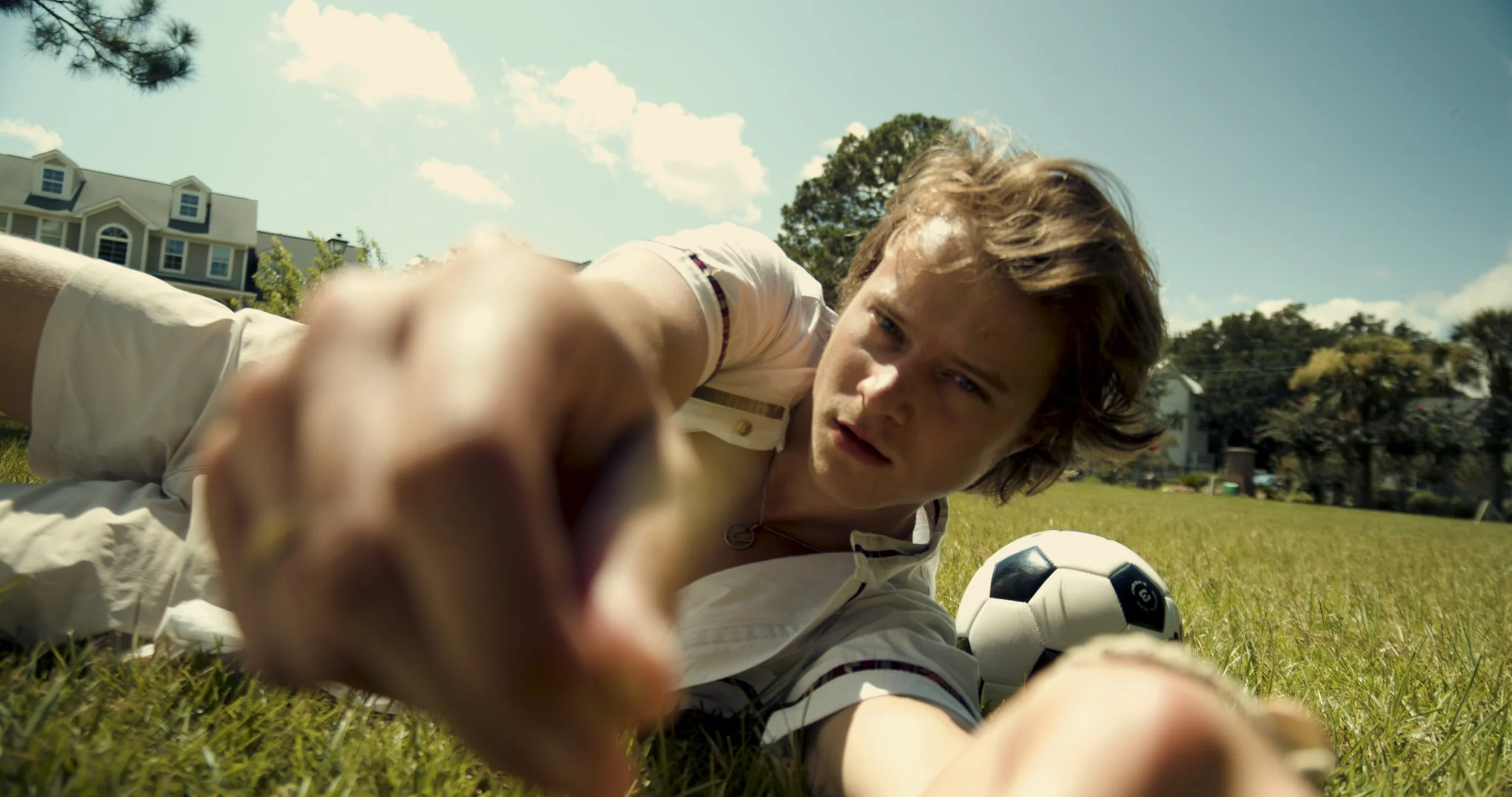 A young man lies on the grass reaching toward the camera with a focused expression, a soccer ball nearby, in a suburban neighborhood on a sunny day.
