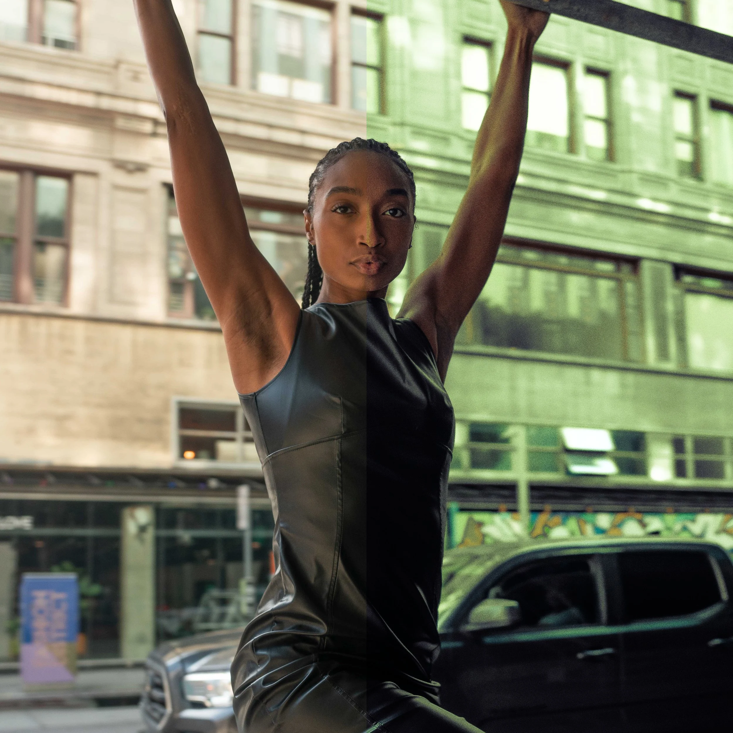 A woman with braided hair and wearing a black sleeveless top, with arms raised, standing in front of a green building with large windows.