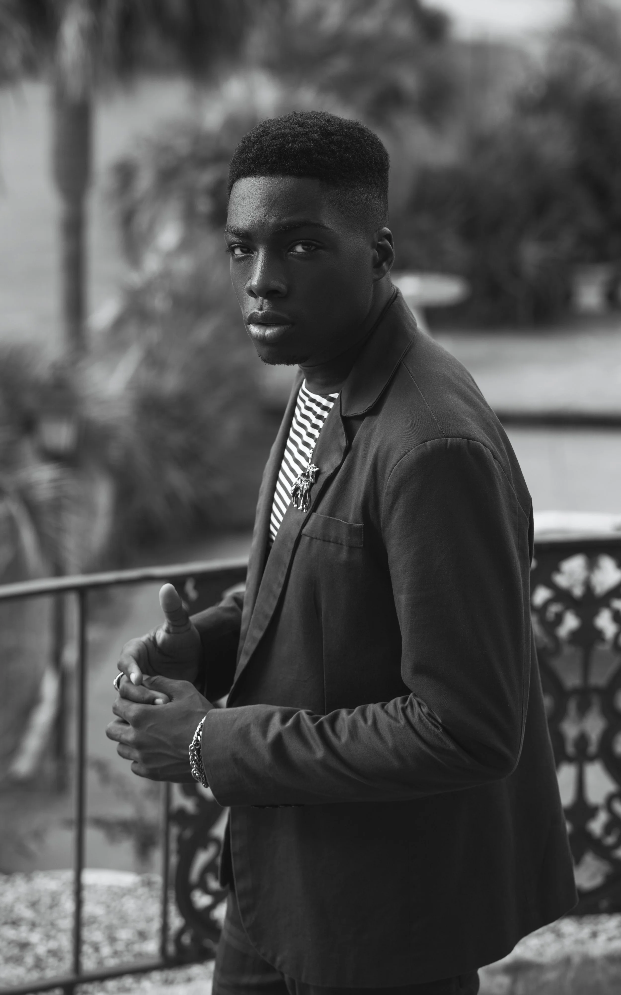 Black and white photo of a young man with short hair, wearing a blazer over a striped shirt, looking directly at the camera with a serious expression outdoors.