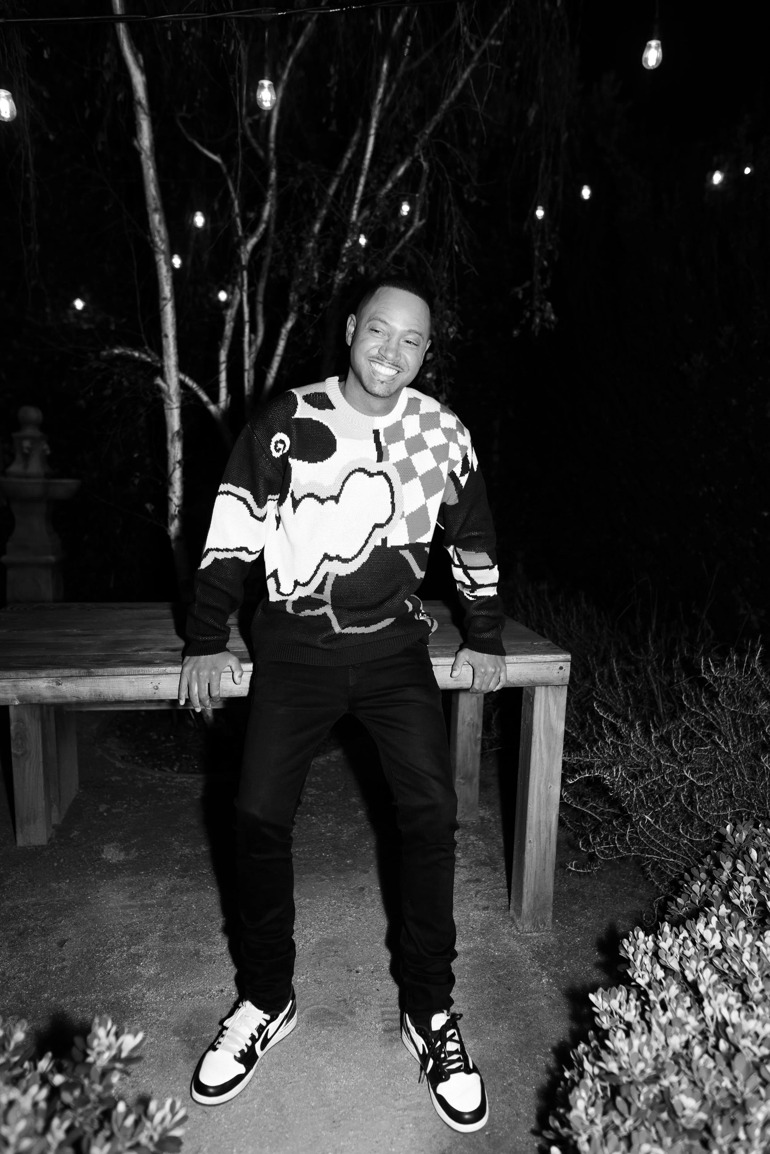 A man smiling and posing outdoors at night, wearing a patterned sweater and sneakers, with trees and string lights in the background.
