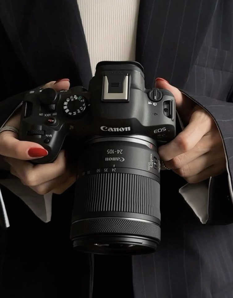 Eine Person hält eine Canon EOS Kamera mit einem 24-105mm Objektiv. Die Person trägt einen schwarzen Anzug mit weißen Streifen. Die Nägel sind rot lackiert.