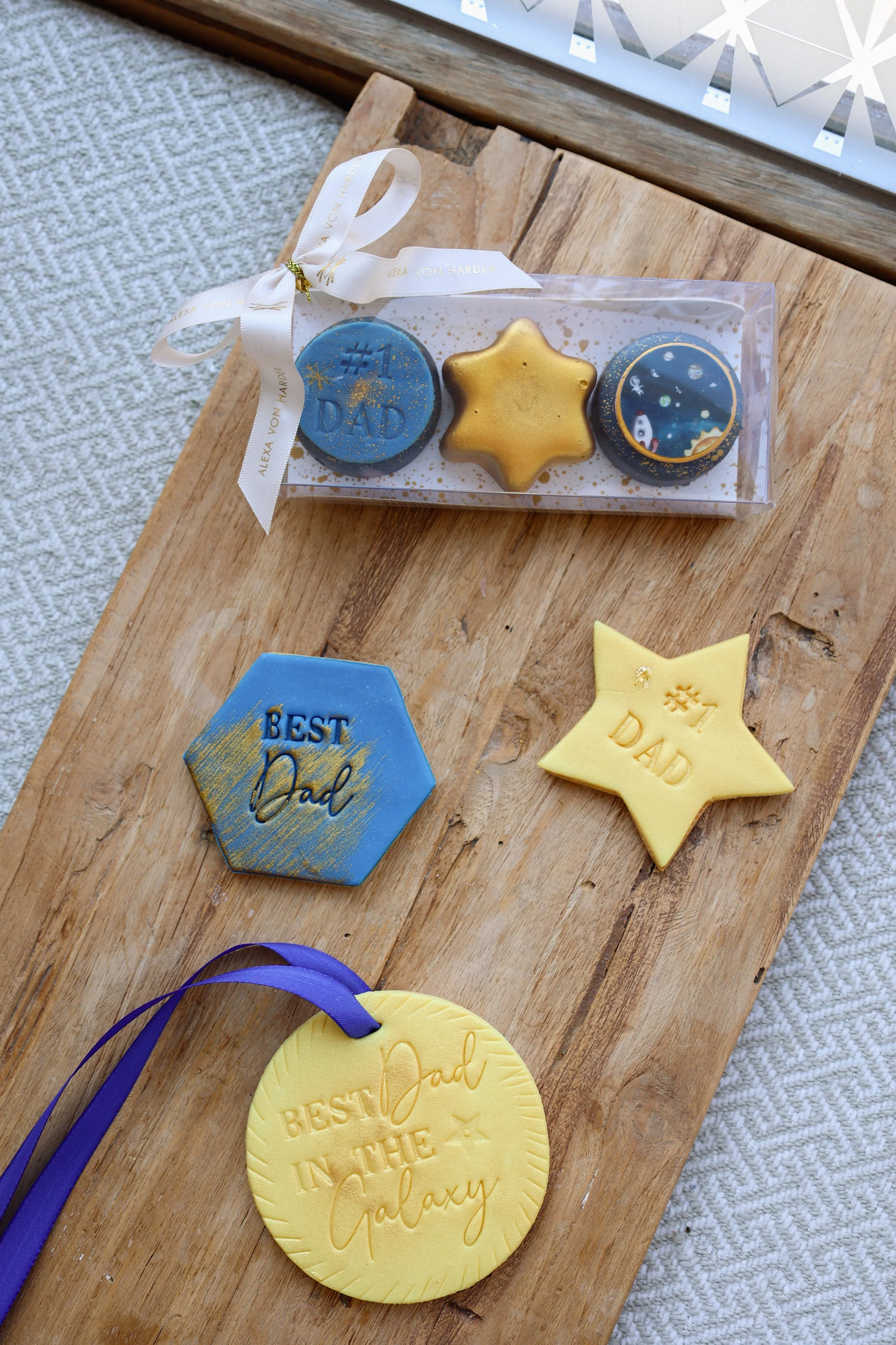 Bunte Weihnachtskekse in verschiedenen Formen und Designs auf einem Holzbrett, verziert mit Glitter und Aufdrucken. Besonders ein Keks mit der Aufschrift "Best Dad in the Galaxy" in Blau und Gold, sowie andere Sterne- und Planetenmotive.