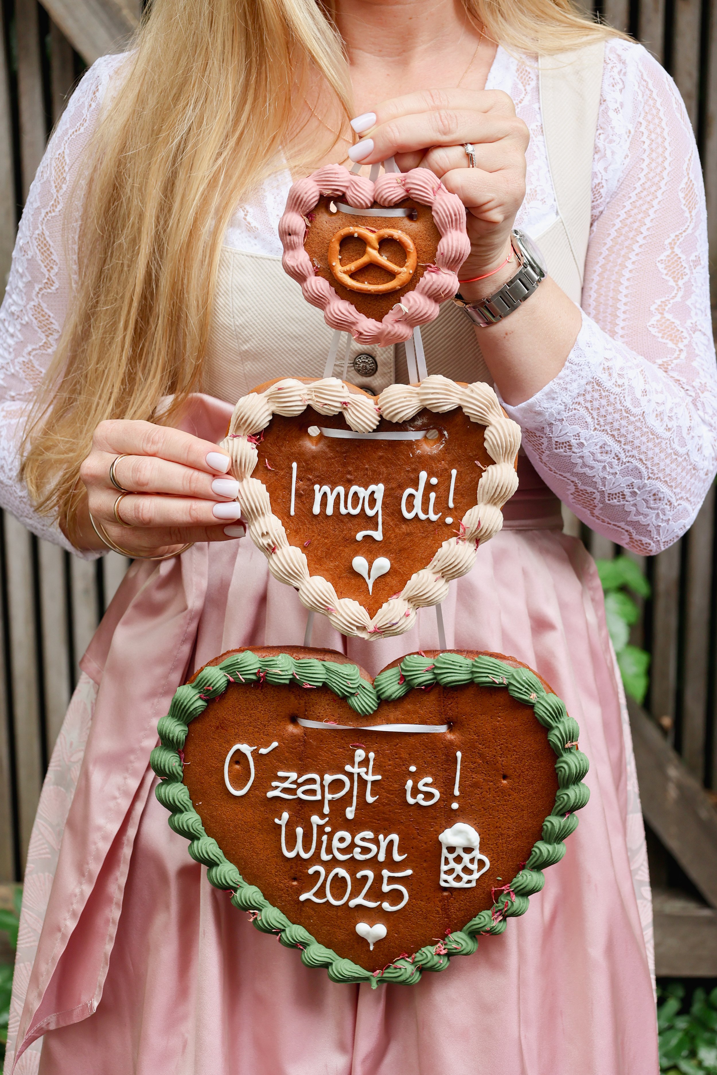 Eine Frau hält eine mit Zuckerguss verzierte Lebkuchenherz mit dem Aufdruck 'O zapft is! Wiesn 2025', begleitet von weiteren Lebkuchenherzen mit ähnlicher Verzierung.