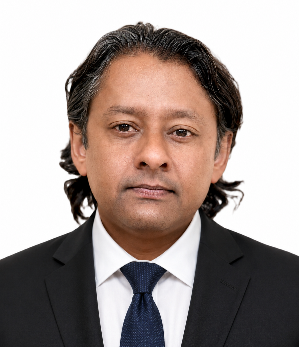 A man with medium-length dark hair, wearing a checked button-down shirt and a dark blue blazer, looks directly at the camera with a neutral expression, against a plain white background.