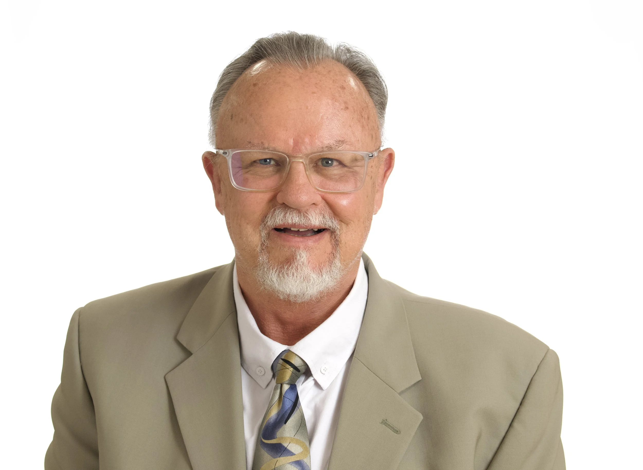 Portrait of an elderly man with gray hair, beard, and glasses, wearing a beige suit, a white shirt, and a multicolored tie, smiling against a white background.