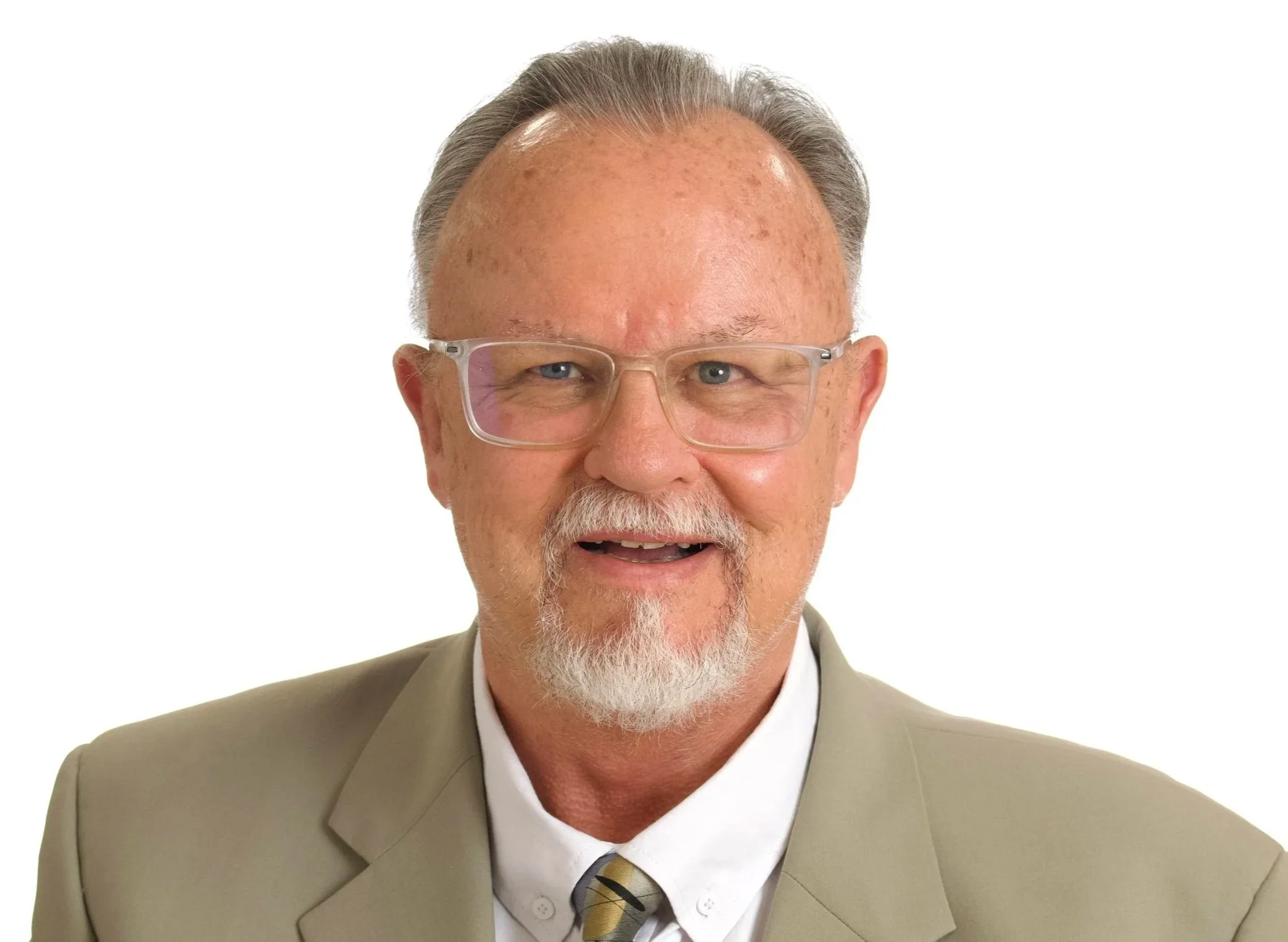 Portrait of an elderly man with gray hair, beard, and glasses, wearing a beige suit, a white shirt, and a multicolored tie, smiling against a white background.